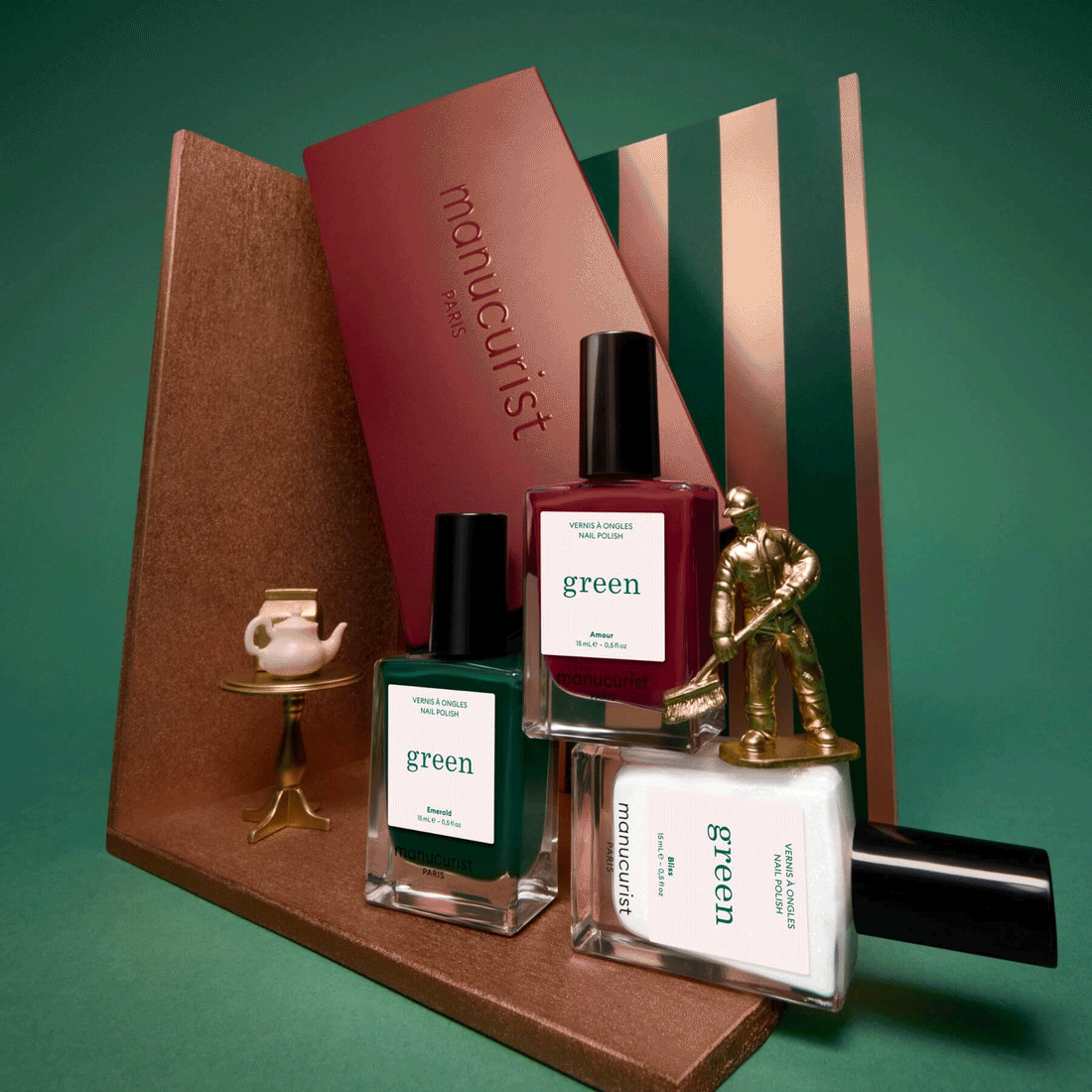 The Manucurist Customisable Trio Green nail polish set—featuring green, red, and white shades—is beautifully displayed with a gold figurine, a tiny teapot on a gold tray, and decorative panels in the background, making it perfect for creating your own unique manicure style.