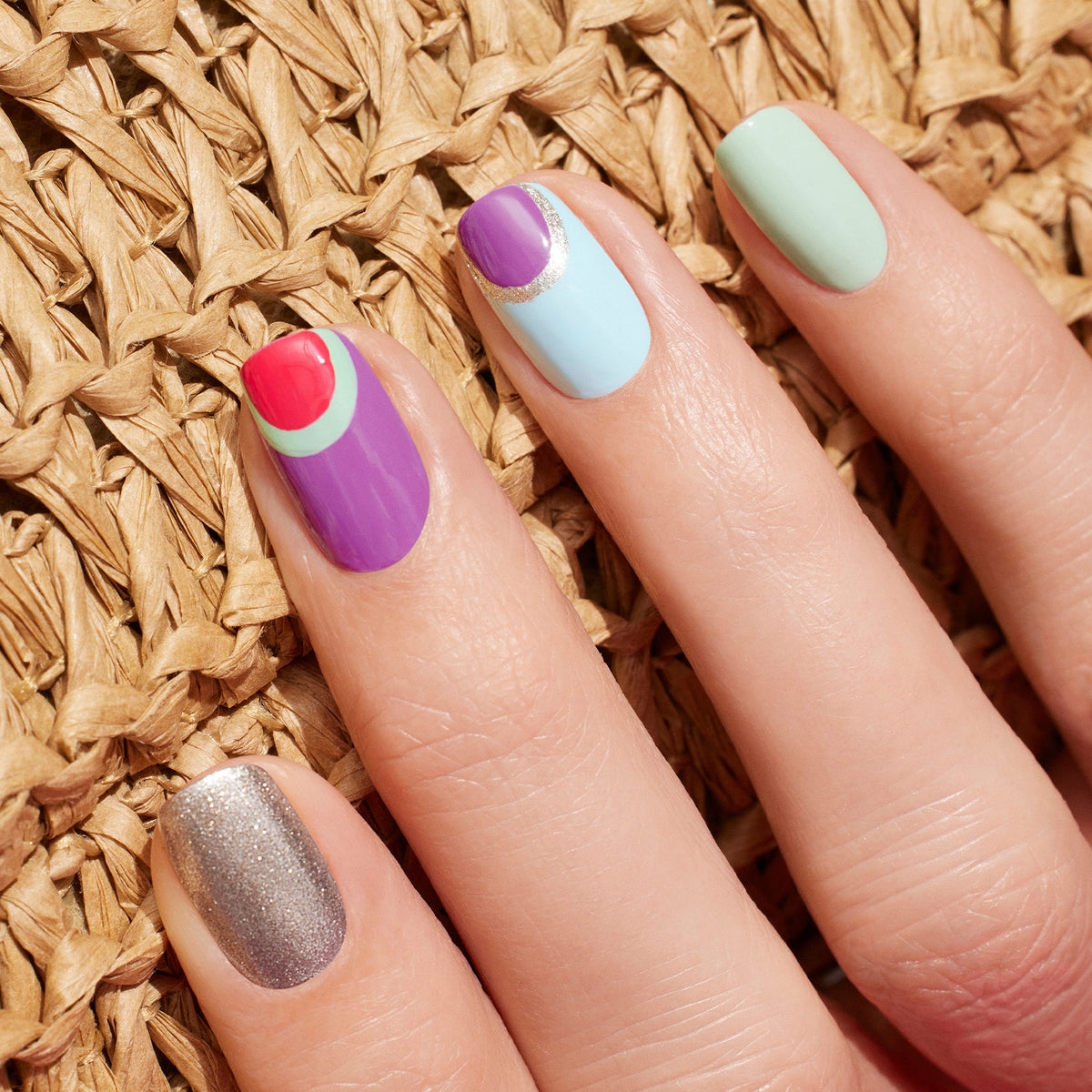 A hand rests on a woven surface, showcasing five nails painted uniquely with Manucurist Violette, silver glitter, light green Green Flash gel polish, blue with a purple half-moon and silver arc, and retro purple with red and green accents.
