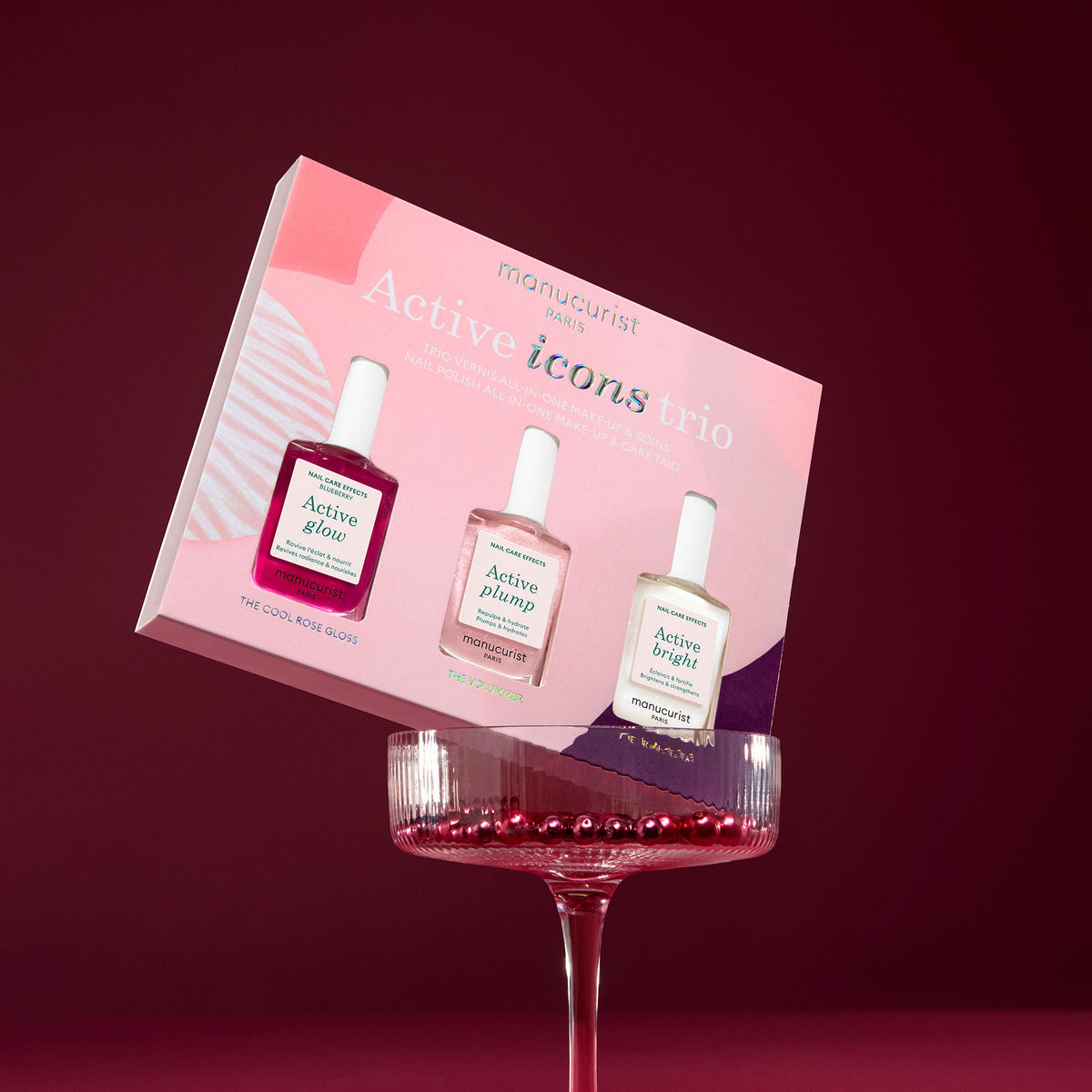 The Manucurist Active Icons Trio, a limited edition nail polish gift set featuring Glow, Plump, and Bright, is balanced on a wine glass of red beads against a pink and burgundy background.