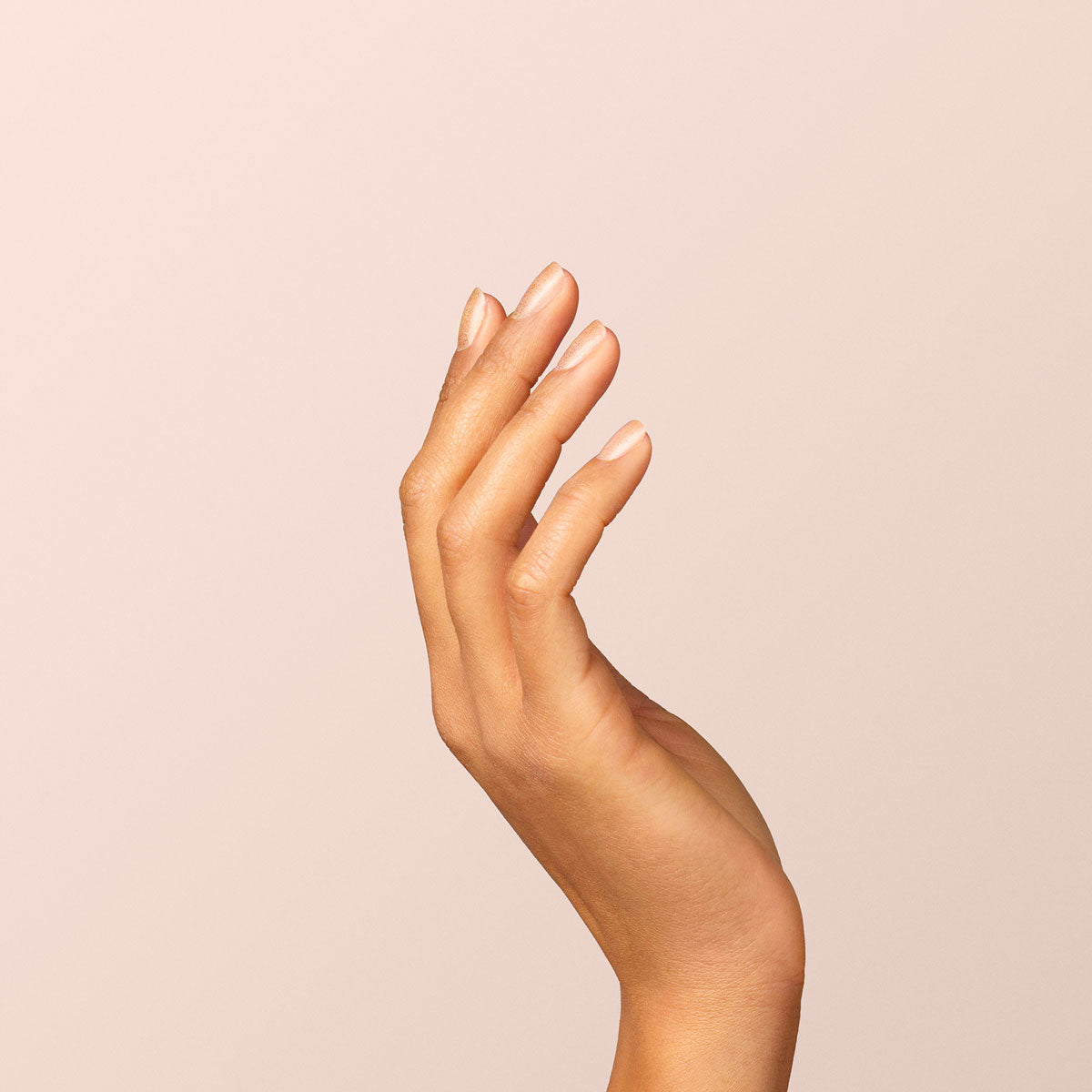 A close-up of light brown skin fingers, neatly trimmed and painted with Manucurist Gold nail polish, posed gracefully against a plain light beige background.