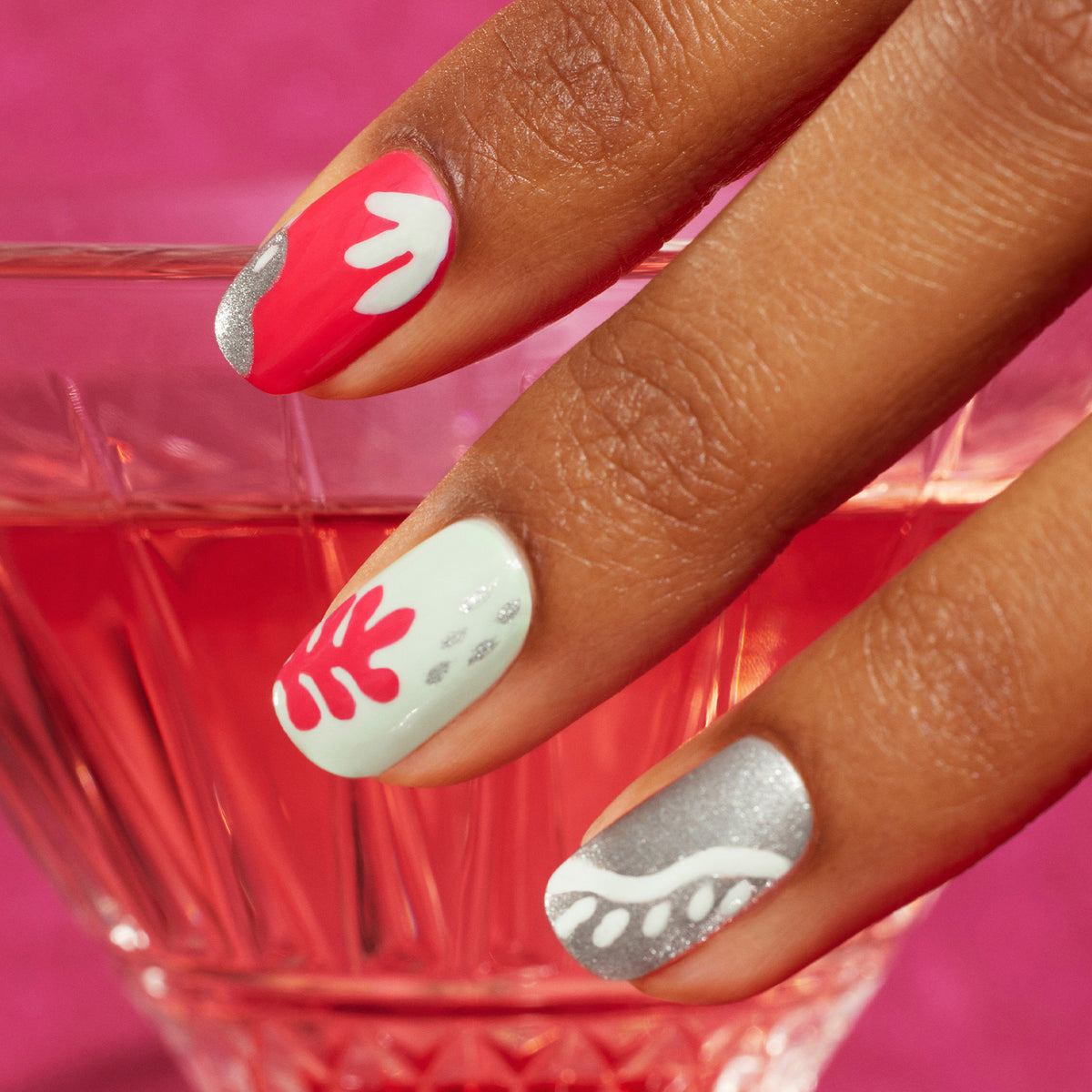 A hand with four fingernails painted in pink, white, silver, and mint green from the Coral reef collection by Manucurist, featuring abstract designs inspired by coral reefs, rests on the rim of a pink glass with a pink drink.