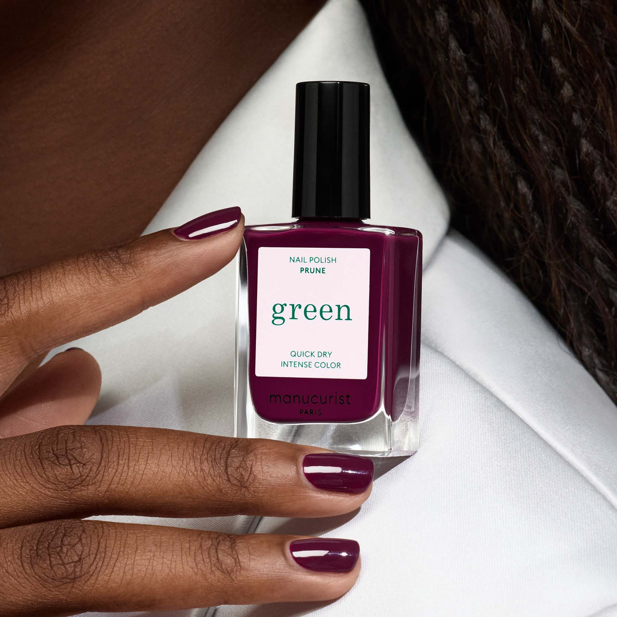 A hand with dark skin holds a bottle of Manucurist "Prune" nail polish; the velvet nails are painted in the same deep purple shade as the polish.