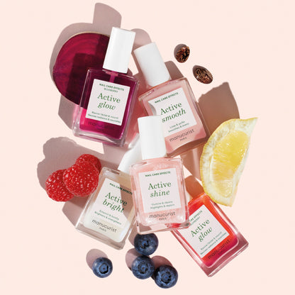 Six bottles from the Active™ Manicure Kit—your manicure essentials—are arranged on a pink background, surrounded by fresh fruit such as raspberries, blueberries, lemon, and beetroot slices to highlight their natural ingredients.