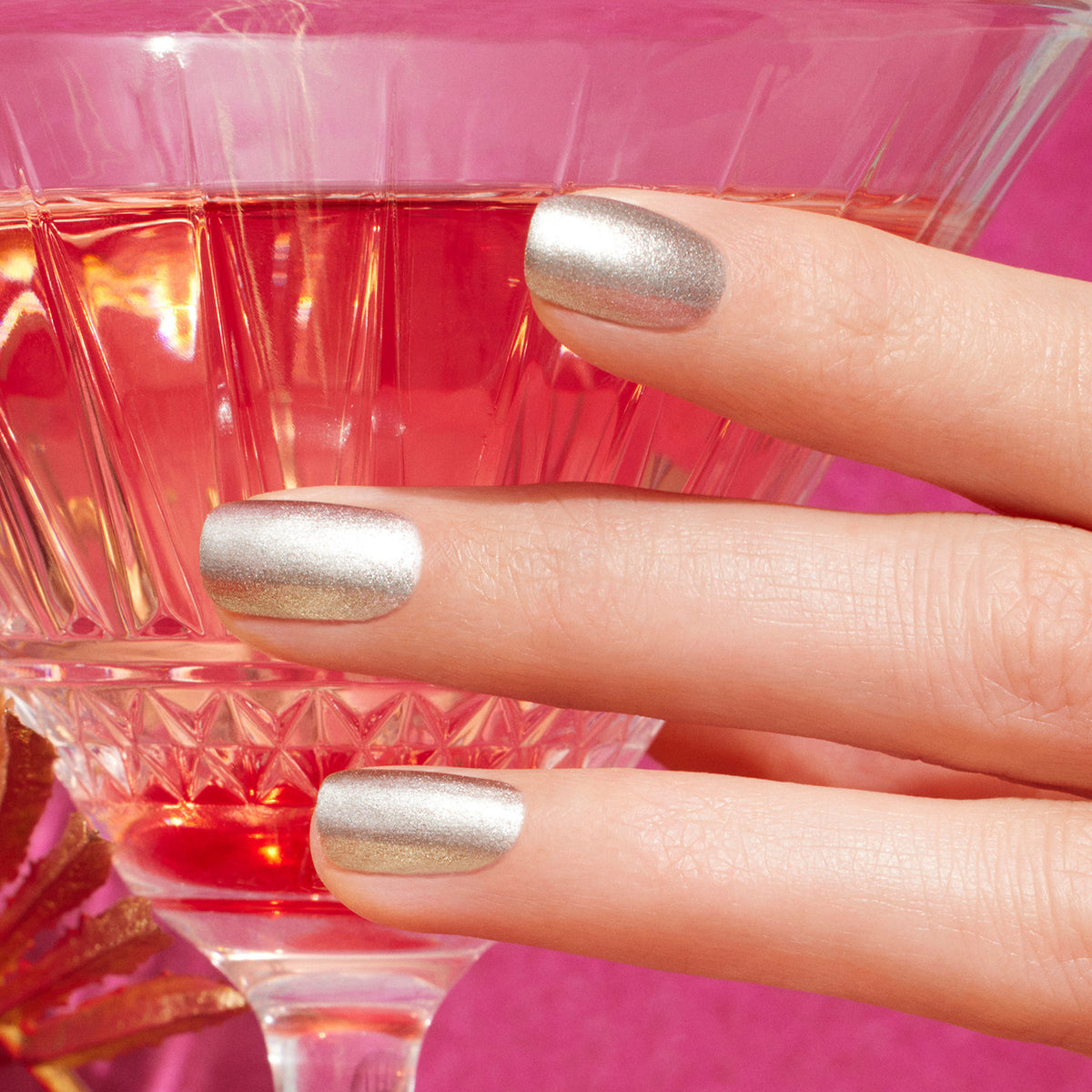 A hand with shimmery silver "Shiny Summer" nail polish by Manucurist from the Summer Love collection holds a pink cocktail glass against a vibrant pink background.