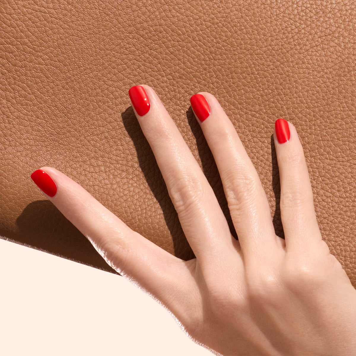 A hand with neatly manicured nails painted in Anémone by Manucurist, a juicy red shade, rests on a textured brown leather surface.