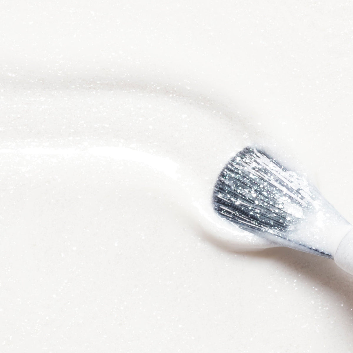 A makeup brush sweeps Manucurist’s Bliss across a smooth, white surface, creating a sparkling, textured effect reminiscent of pearly white gel polish.