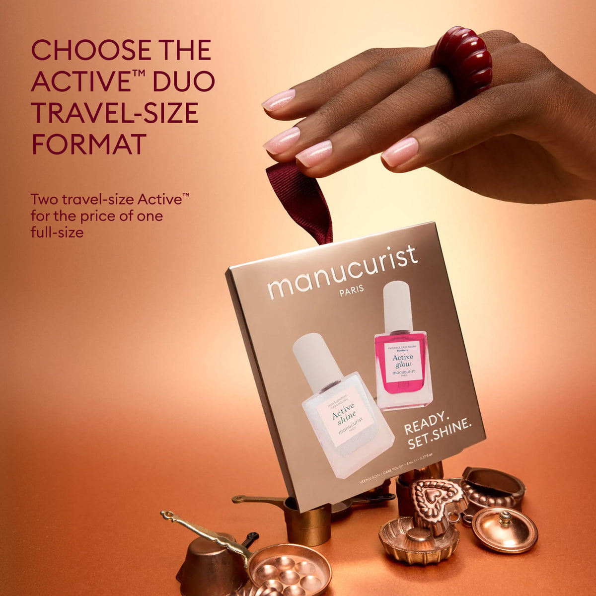 A hand adorned with a red ring holds a ribbon fastened to a Manucurist Active™ Shine Icy nail polish duo box, which features illuminating formulas for brighter-looking nails. Arranged beneath soft lighting on a brown surface are miniature bowls and cups.