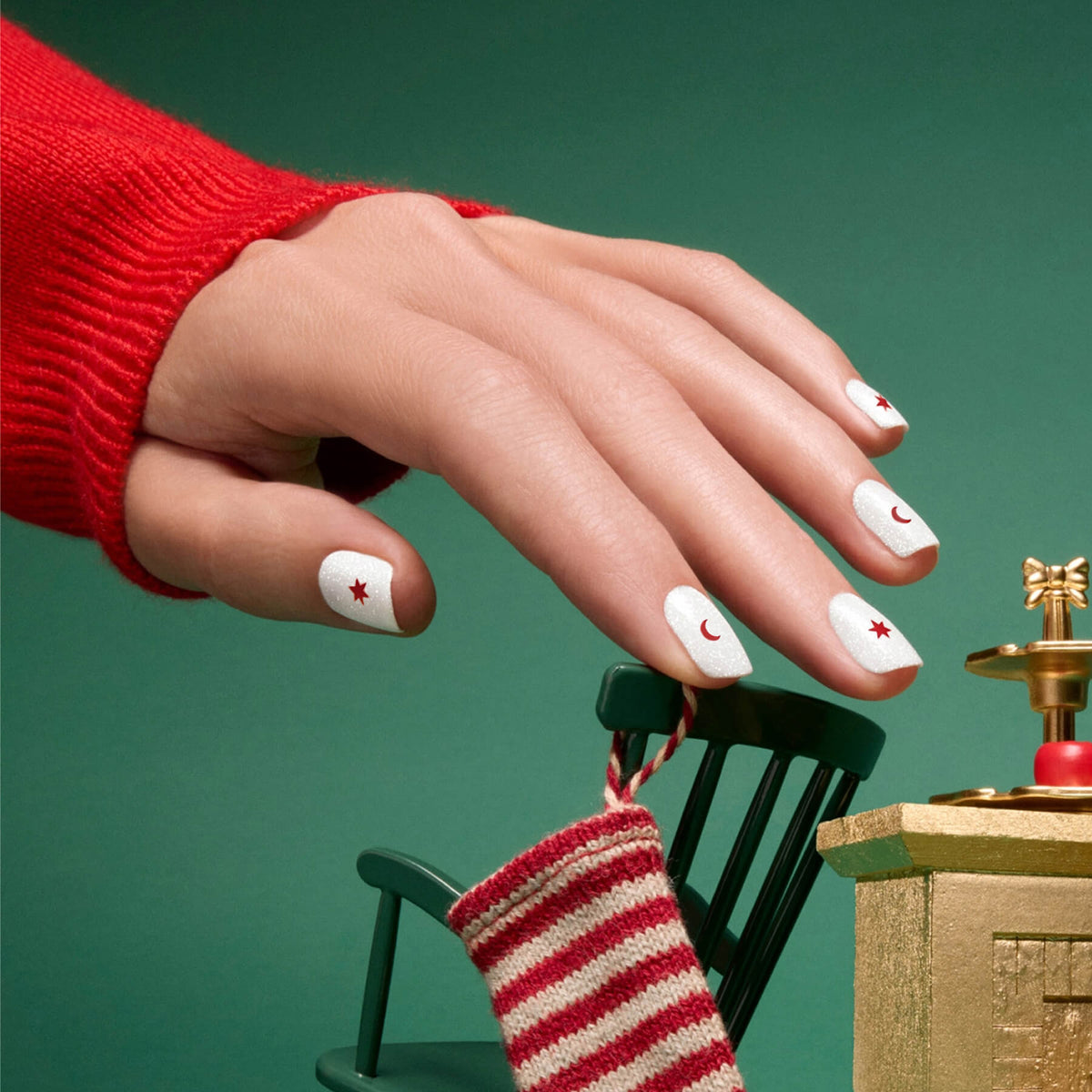 A hand featuring a festive manicure—white nails decorated with red crescent moons and stars, likely achieved with Manucurist Stickers Nail Art Holidays—reaches toward a small striped Christmas stocking resting on a green rocking chair, all set against a green background.