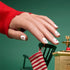 A hand featuring a festive manicure—white nails decorated with red crescent moons and stars, likely achieved with Manucurist Stickers Nail Art Holidays—reaches toward a small striped Christmas stocking resting on a green rocking chair, all set against a green background.