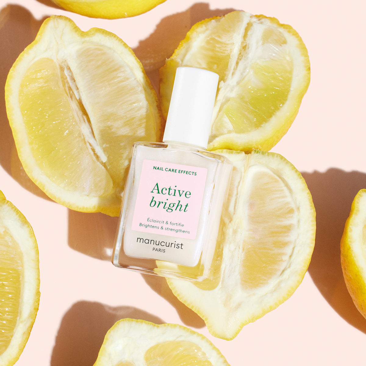 A bottle of Manucurist Active™ Bright is surrounded by halved yellow lemons on a light peach background, highlighting this bio-based nail care product as the ideal option for naturally vibrant and healthy-looking nails.