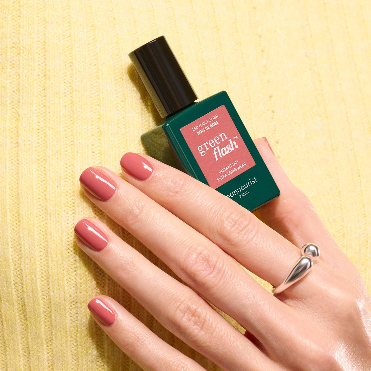 A hand with neatly manicured Bois de rose pink nails holds a Manucurist Green Flash gel polish bottle against a yellow textured background, wearing a modern silver ring.