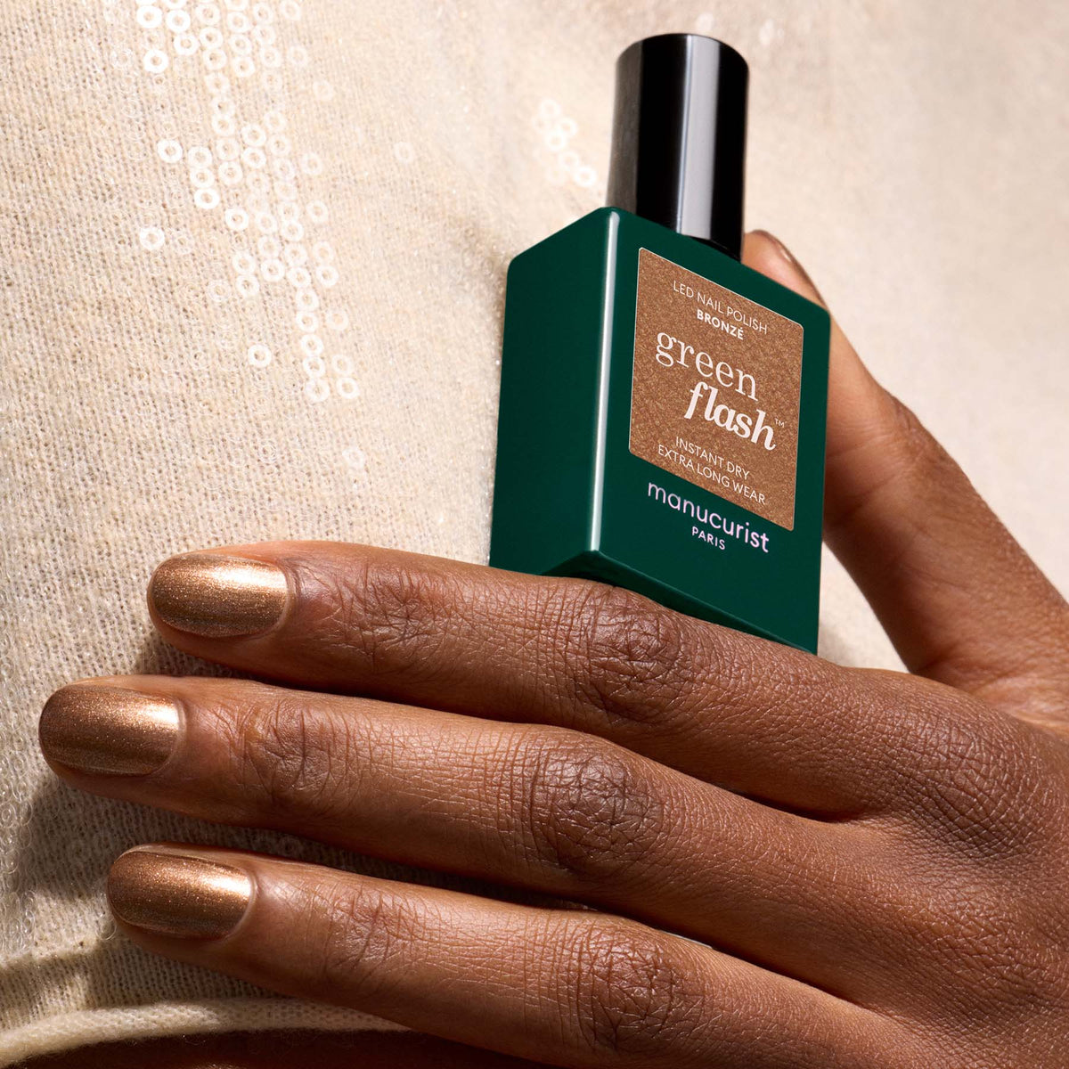 A hand with metallic bronze nails holds a bottle of Manucurist Bronzé polish against a beige sequined fabric background.