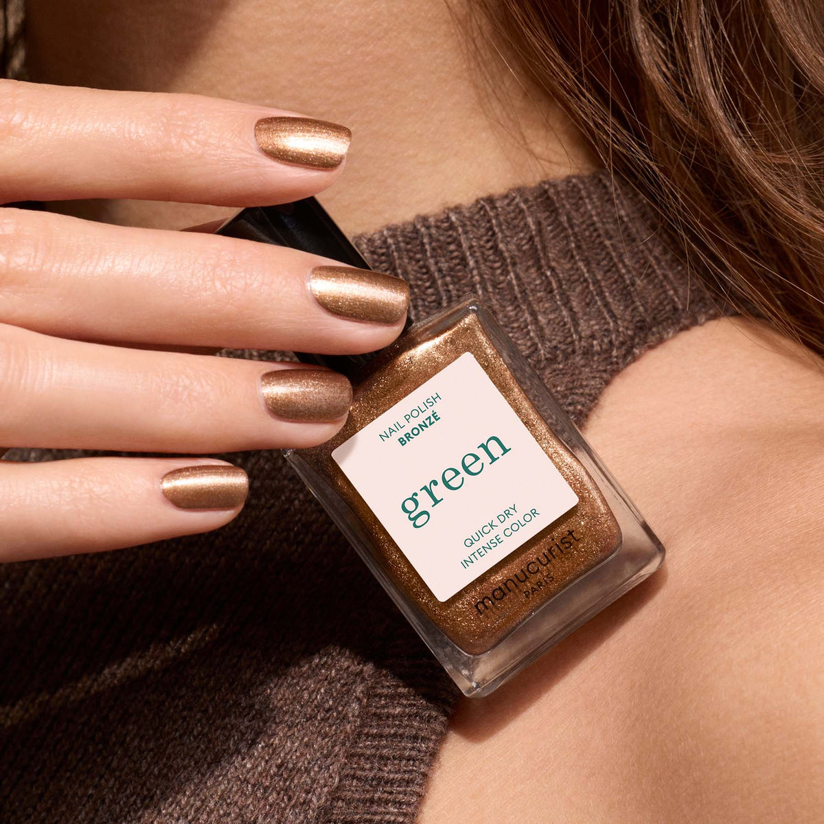 A hand with Bronzé copper nail polish from Manucurist holds a matching bottle labeled Manucurist, set against a brown ribbed sweater.