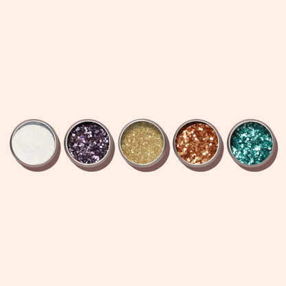 Five small round tins from Manucurist, featuring lilac biodegradable glitter made from plant cellulose plus white, purple, gold, copper, and teal shades, are arranged in a row—perfect for eco-friendly nail art.