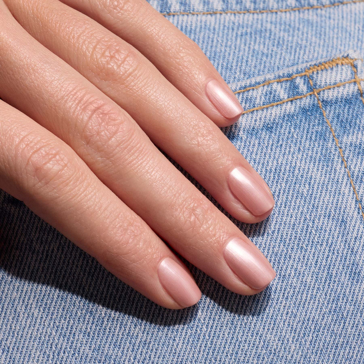 A close-up of a hand with neatly manicured nails painted in Carnation by Manucurist, a pearly pink beige polish from the Green™ range, resting on light blue denim fabric.