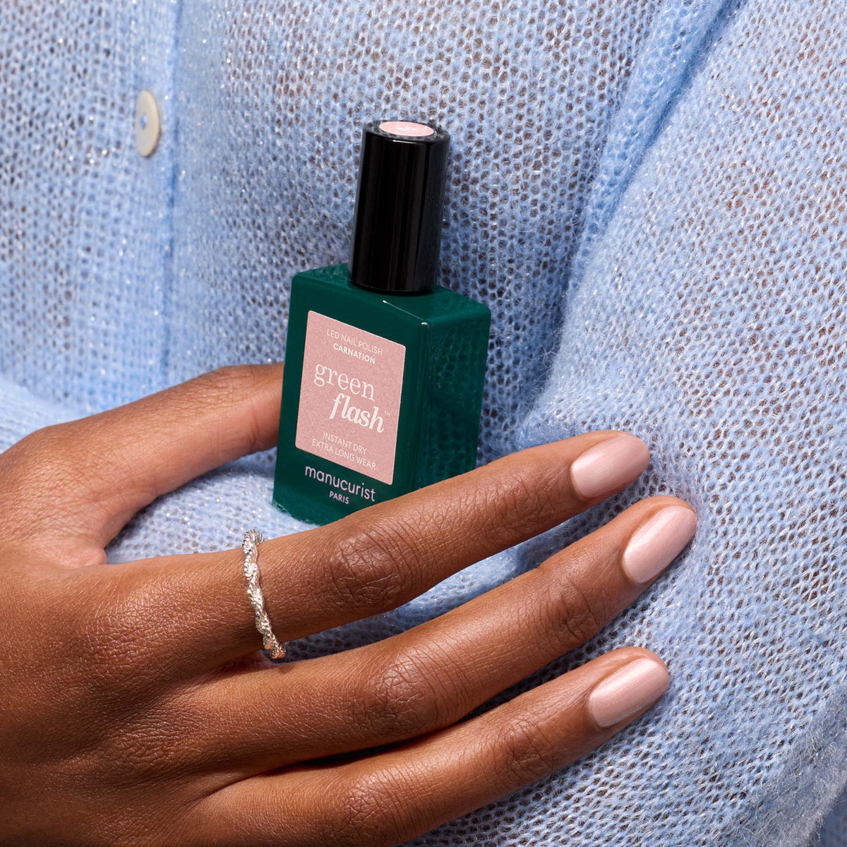A hand with pearly pink beige nail polish and a silver ring rests on a light blue knit sweater, holding a bottle of Manucurist Carnation gel polish.