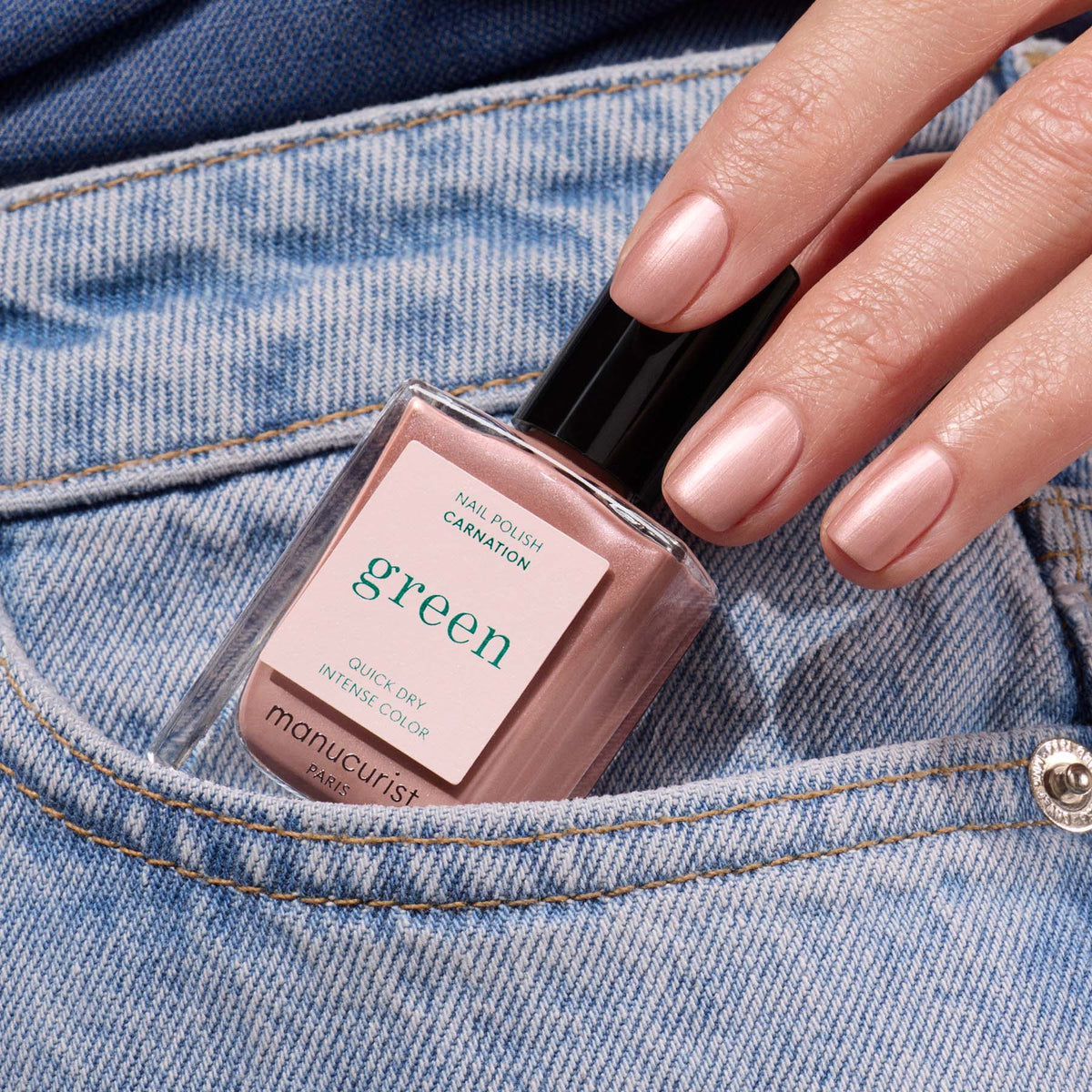 A hand with nude pink nails holds a Manucurist Carnation polish bottle, resting it in the front pocket of blue jeans.
