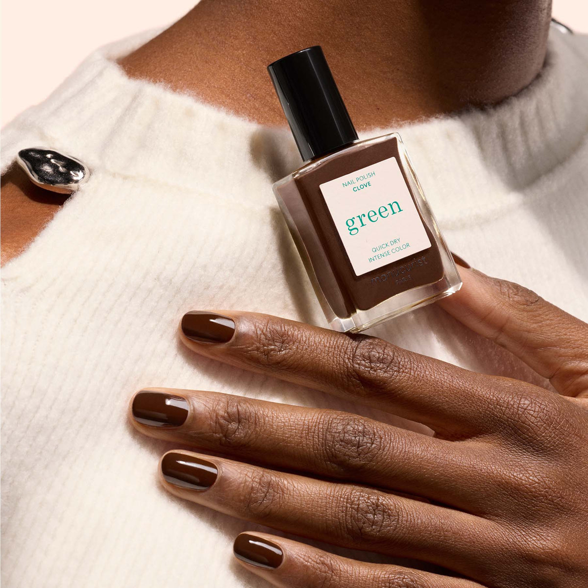 A person with medium-dark skin tone wears Clove nail polish from Manucurist, holding a matching “green” labeled bottle against a cream sweater with a large black button.