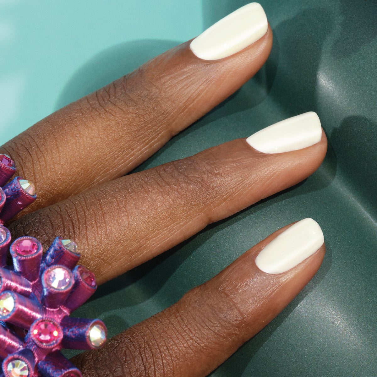 A close-up of a hand with glossy, off-white Porcelaine polish by Manucurist, wearing a large purple jeweled ring. The background features soft teal and dark green surfaces.