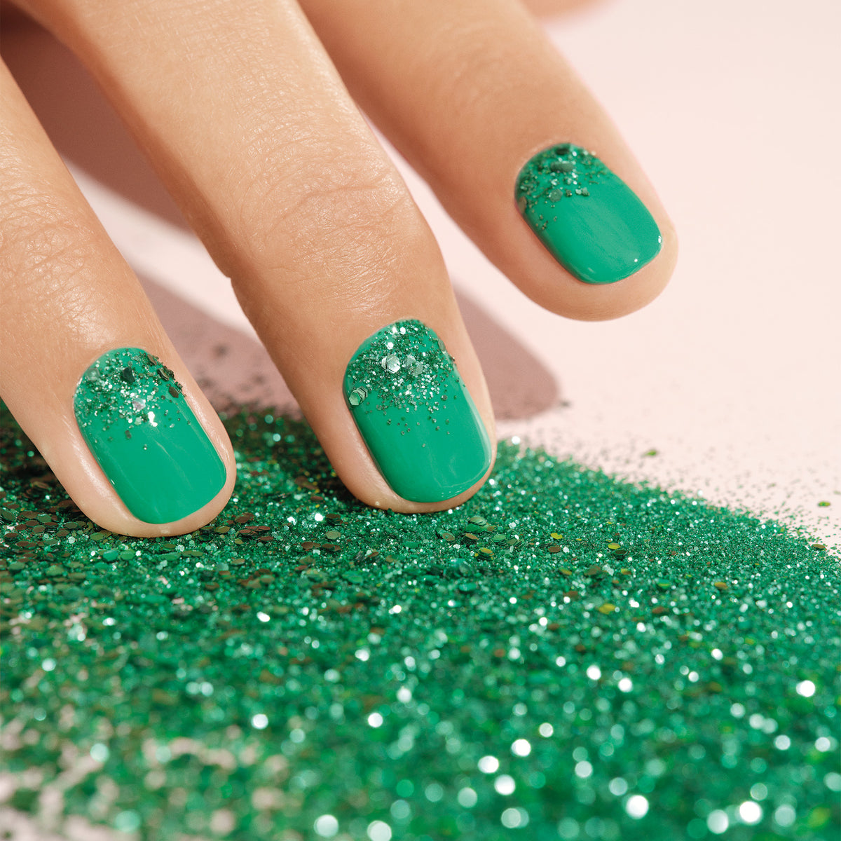 A hand with green nail polish and Manucurist’s Cactus biodegradable glitter on the tips touches a surface sprinkled with the eco-friendly glitter, showcasing sustainable nail art.