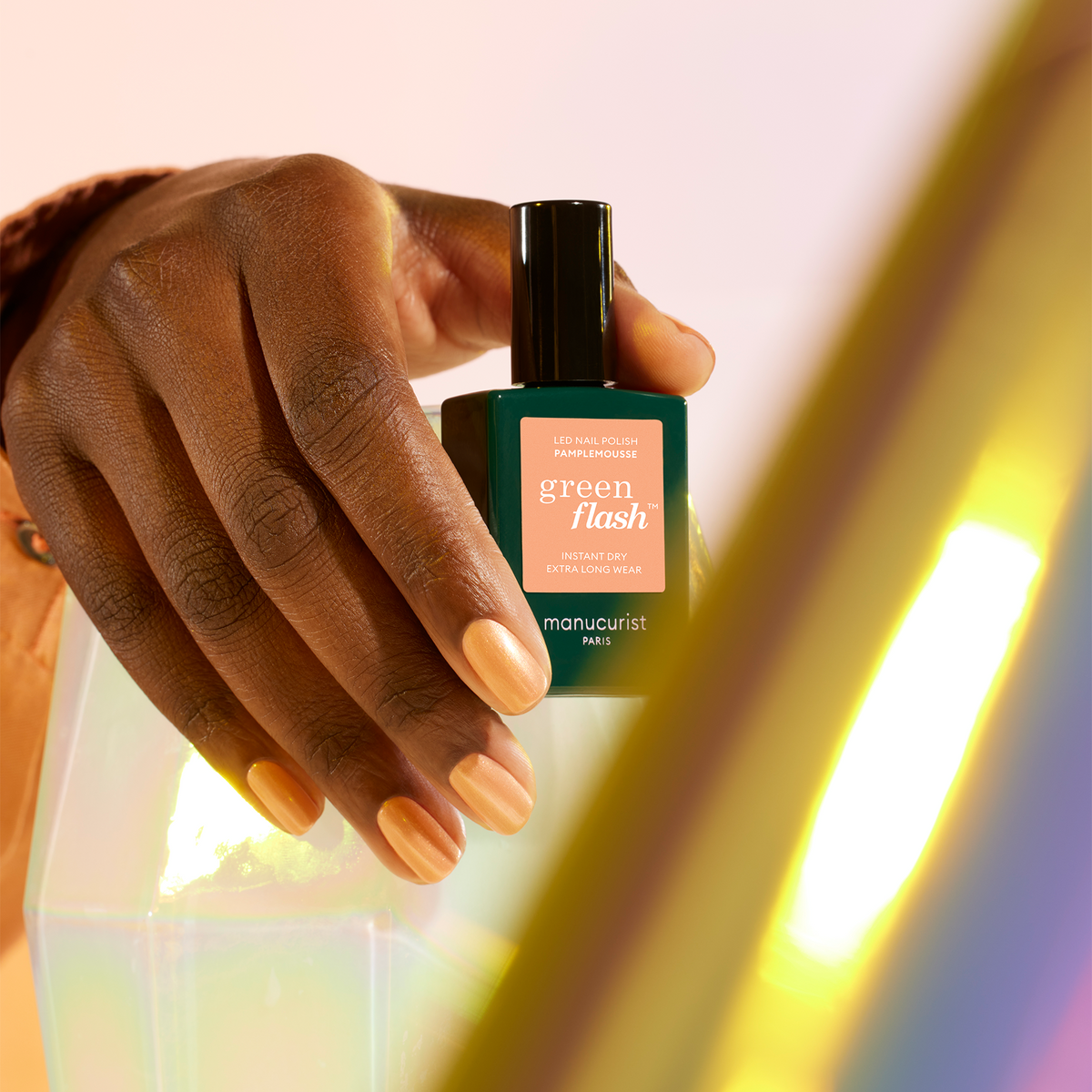 A hand with neatly manicured, natural nails holds a bottle of Manucurist Pamplemousse gel polish against a soft, pastel background with a blurred prism effect in the foreground.