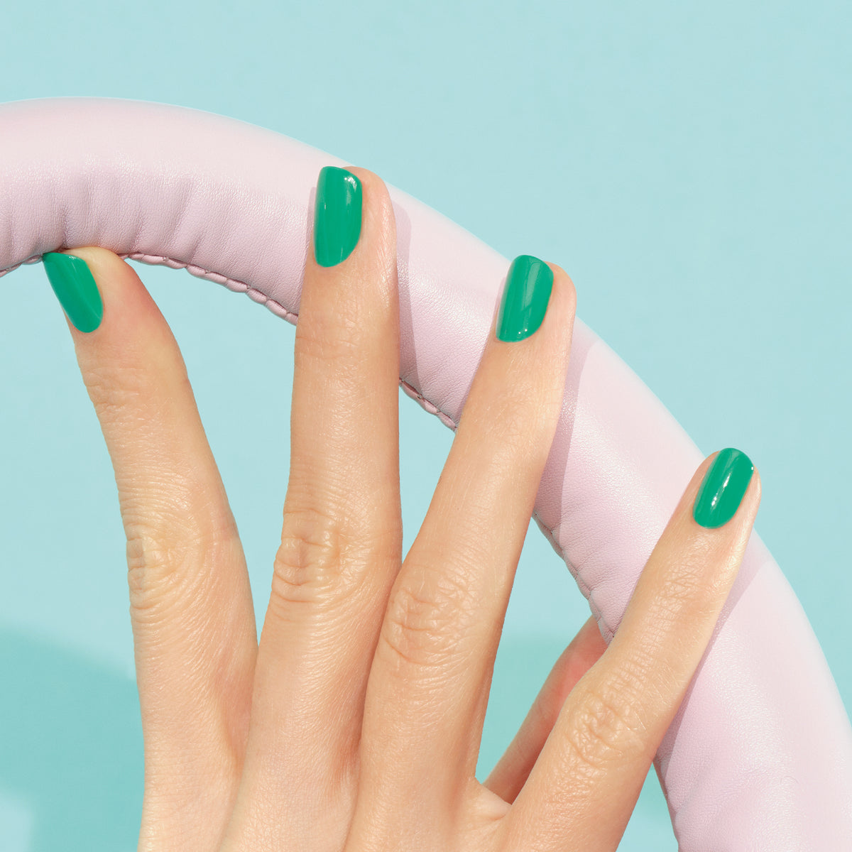 A hand with neatly manicured nails painted in Brazil by Manucurist (electric green) holds a light pink, padded circular object against a light blue background.