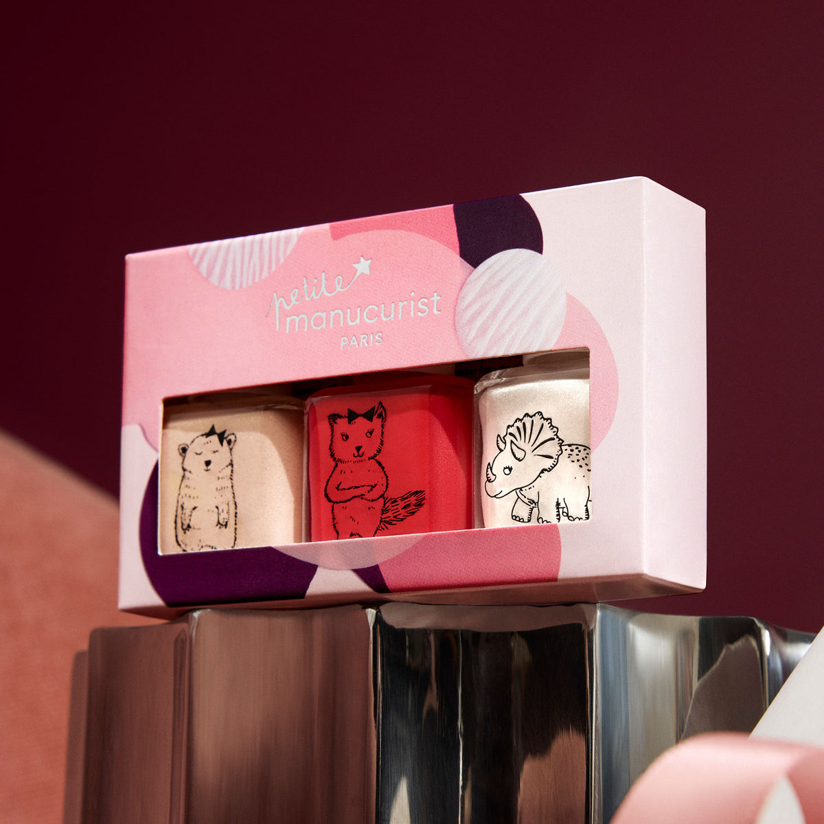 The Manucurist Petite Manucurist Trio is a pink boxed set of three children’s nail polishes made from natural origin, each with animal illustrations: a dog on beige, a fox on red, and a triceratops on white.