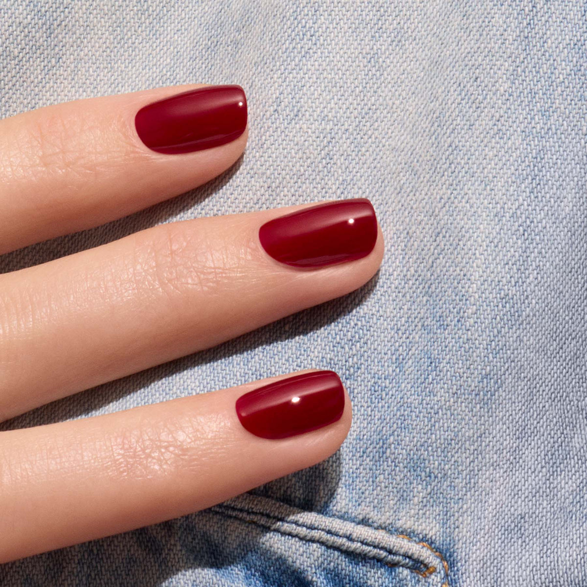 A close-up of a hand with neatly manicured nails painted in Manucurist’s Dark Pansy, a glossy velvet red shade, resting on light blue denim fabric.