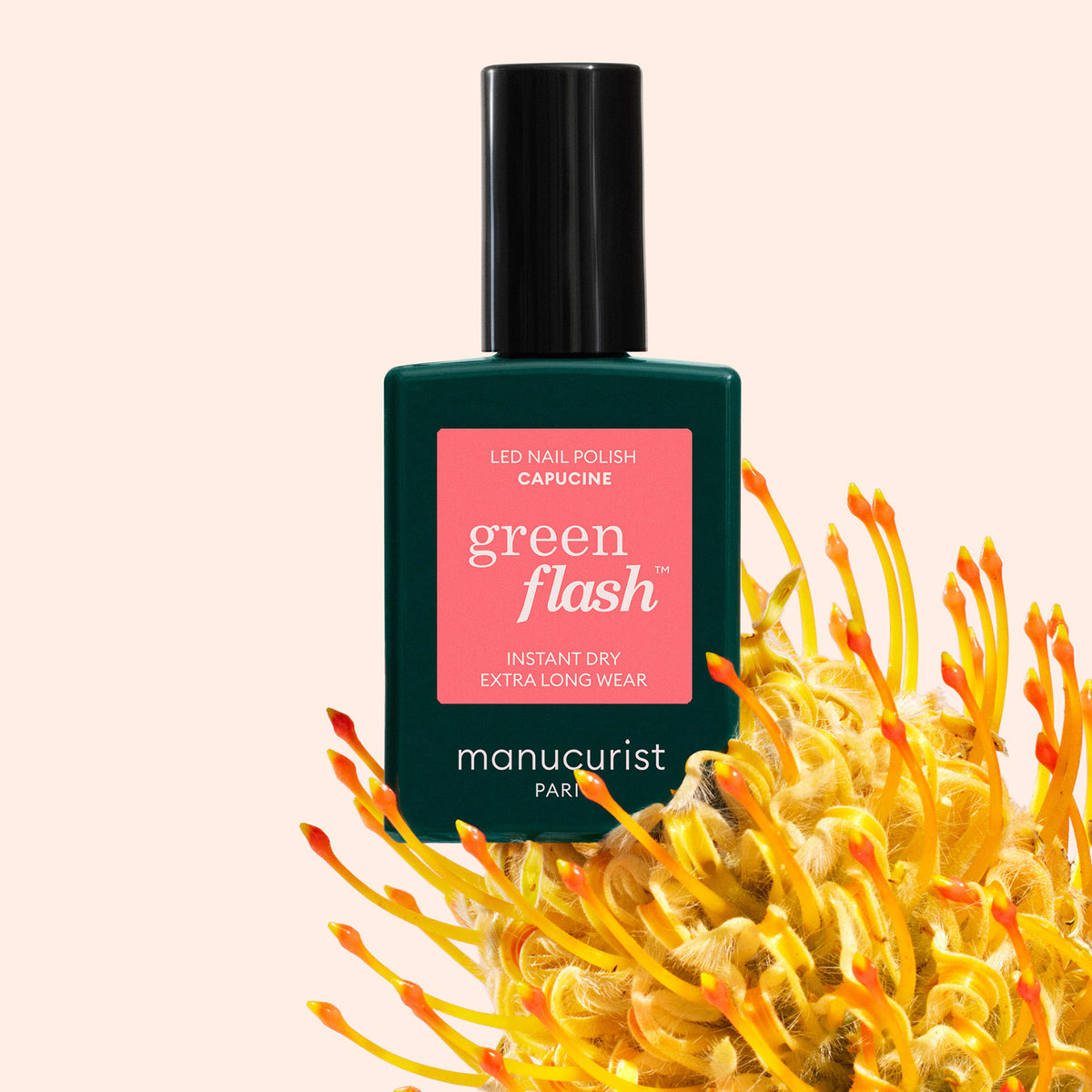A bottle of Manucurist Capucine Green Flash gel polish stands upright, partially surrounded by a vibrant yellow-orange flower, against a light peach background.
