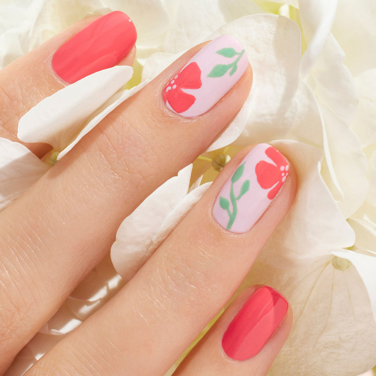 A close-up of a hand with manicured nails: three nails are painted with Manucurist Capucine gel polish, while two feature floral designs with red flowers and green leaves on a pale pink base, resting on white hydrangea petals.