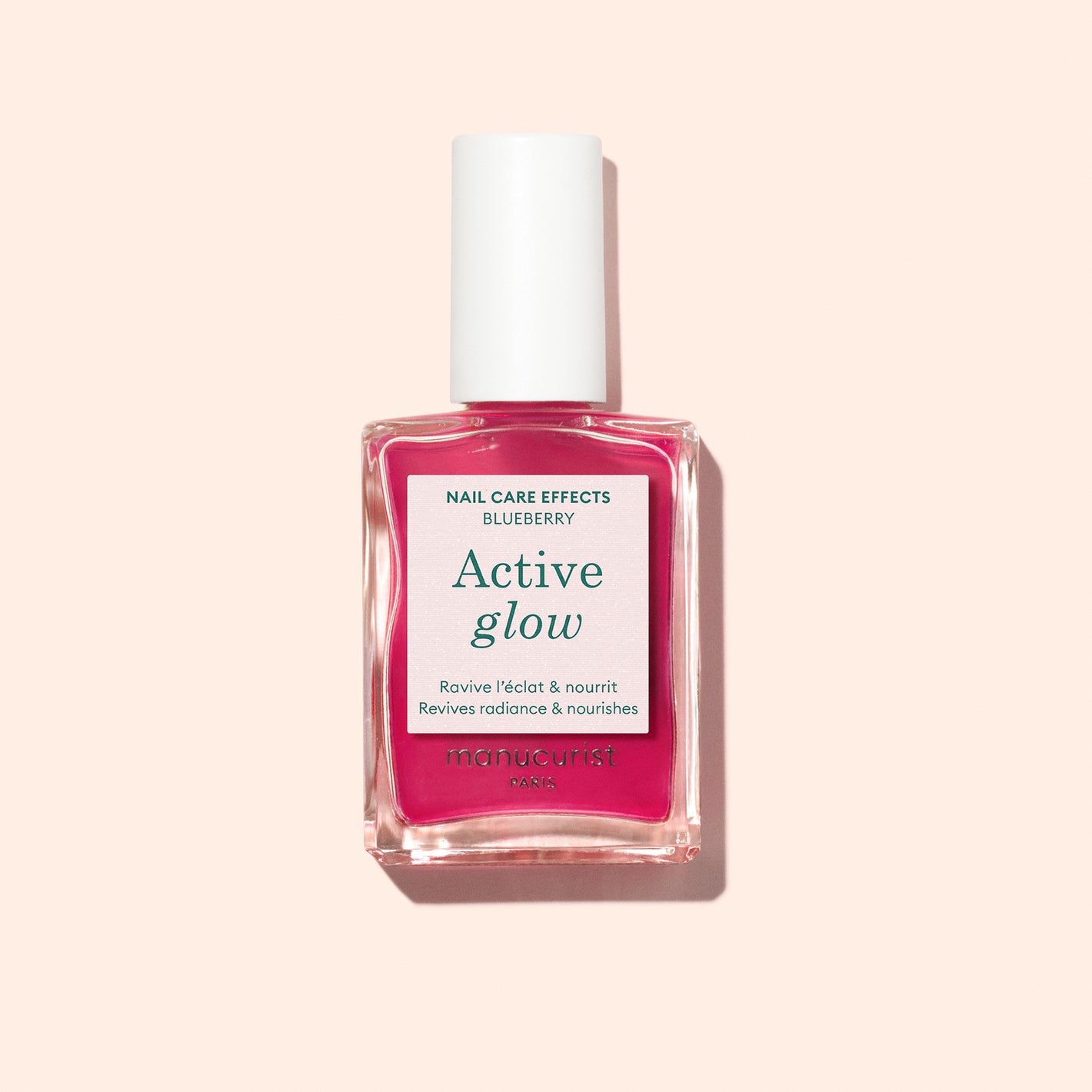 A bottle of Manucurist Active Glow Blueberry nail polish, a bright pink shade with a white cap, sits on a pale peach background—ideal for radiant shine and expert nail care.