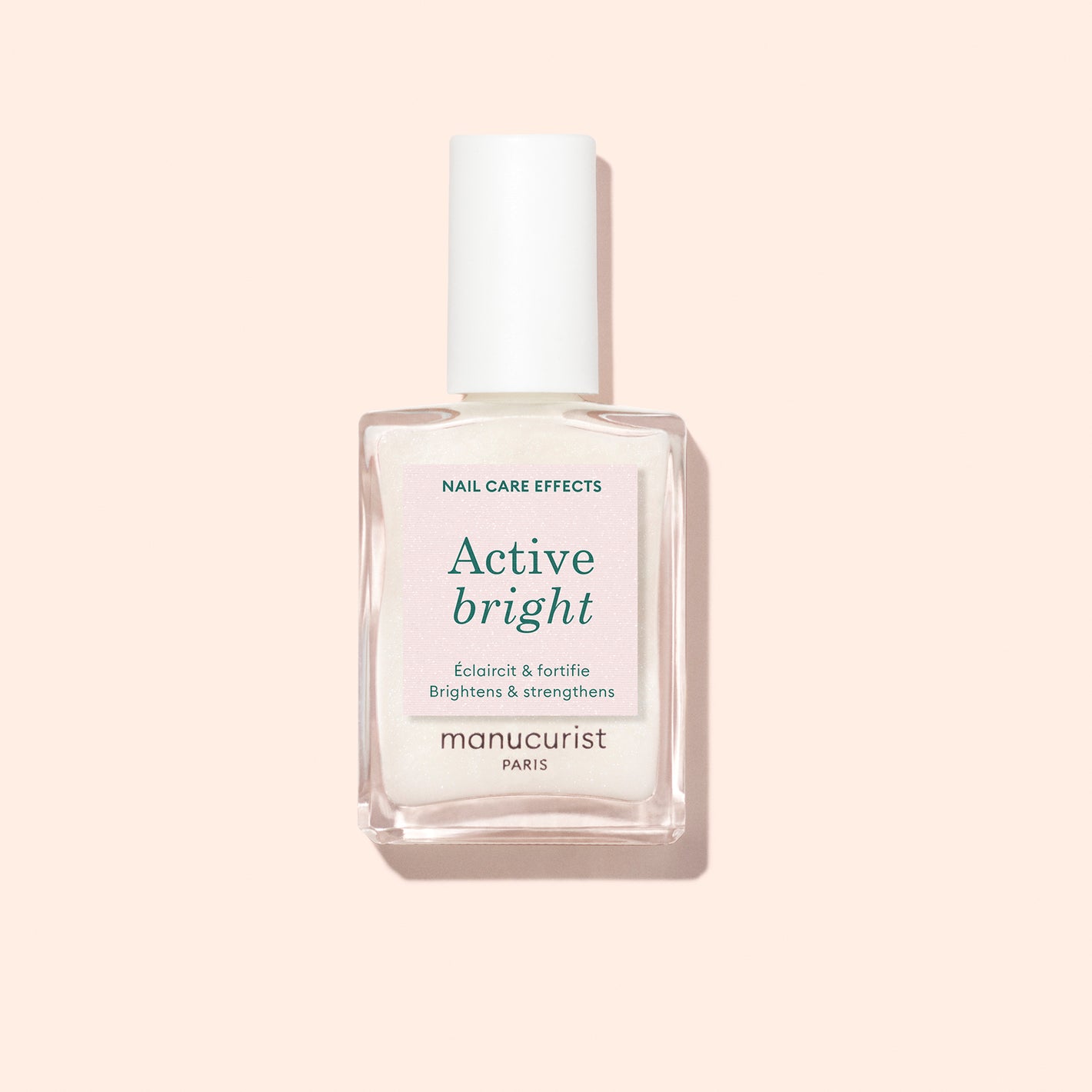 A clear bottle of Manucurist Active Bright nail polish with a white cap is set against a pale pink background. The label showcases its nail care benefits, such as brightening and strengthening.