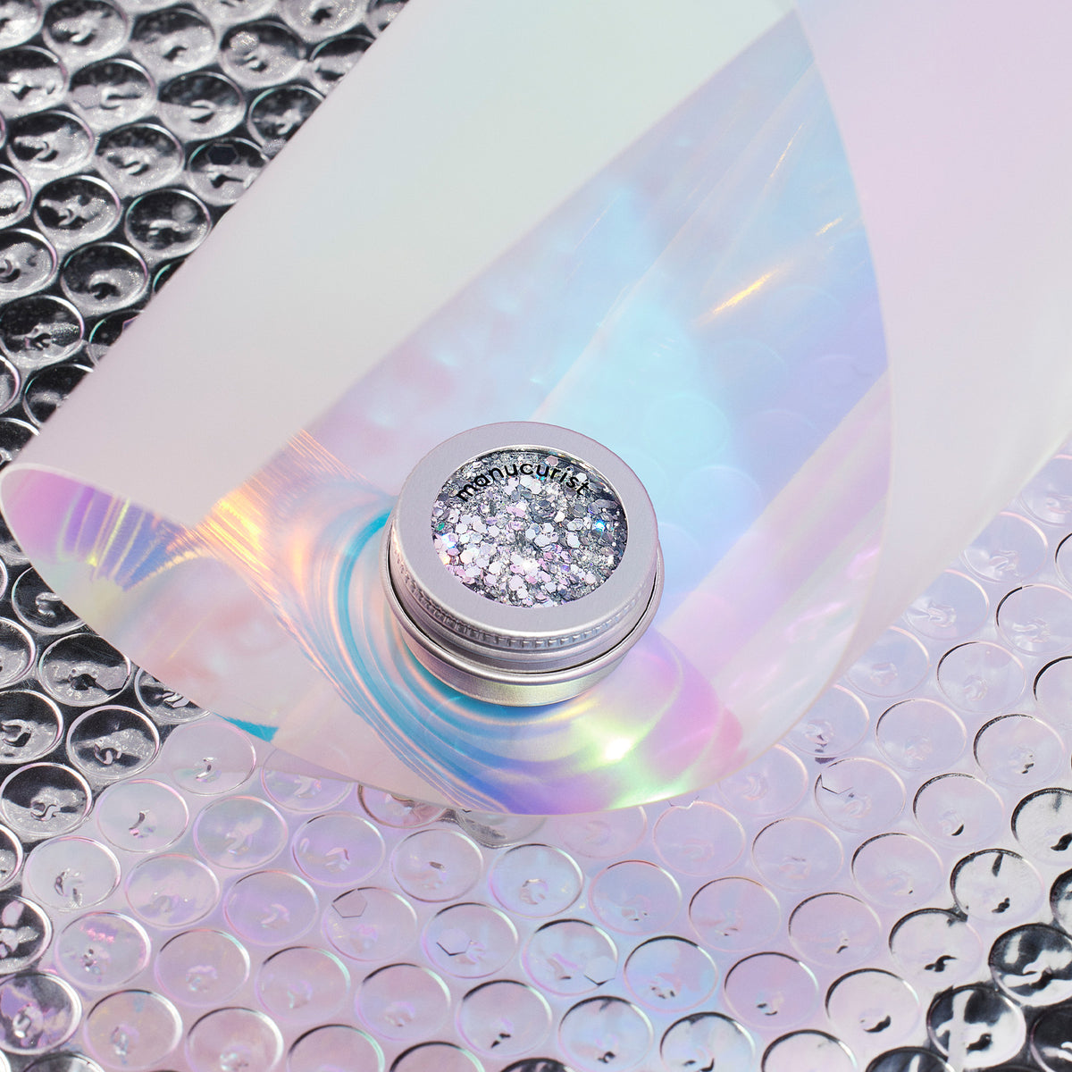 A small jar of Manucurist Holo biodegradable glitter sits on iridescent plastic and clear bubble wrap, reflecting pastel pink, blue, and green hues. The label on the lid reads Manucurist.