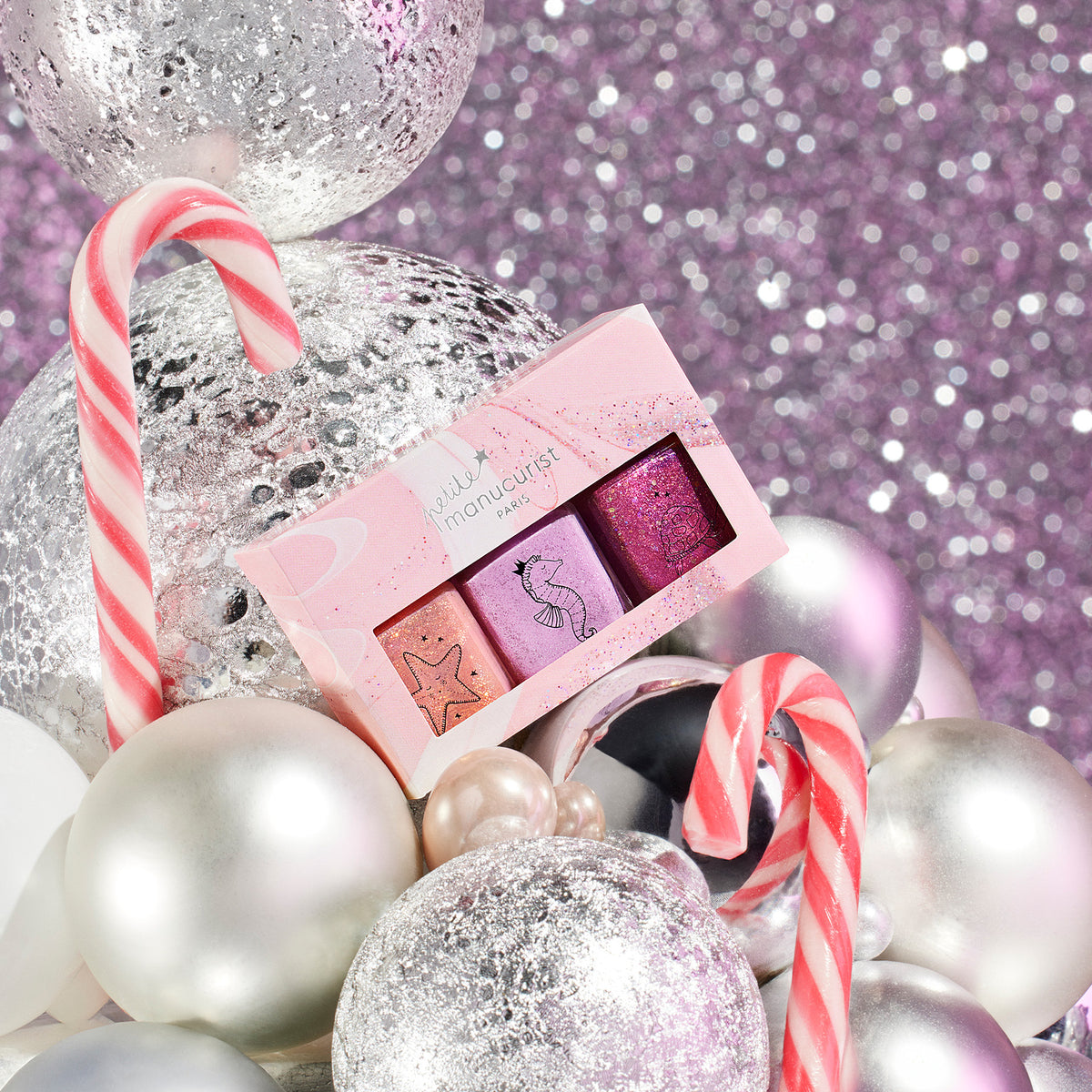 The Manucurist Petite Manucurist Trio, featuring three nail polish colors, is displayed among silver and white ornaments and candy canes on a sparkly pink background, creating a delightful holiday vibe.