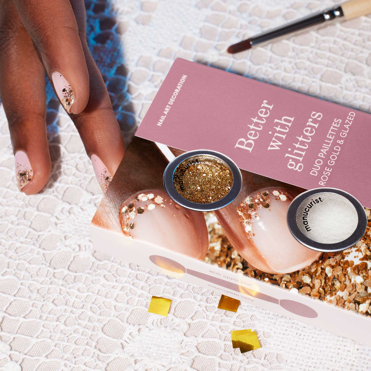 A hand with glittery nude nails rests on lace beside a Manucurist Glitter Duo Rose Gold  Glazed box, highlighting its pearlescent shine. A makeup brush and yellow pearls complete the elegant scene.