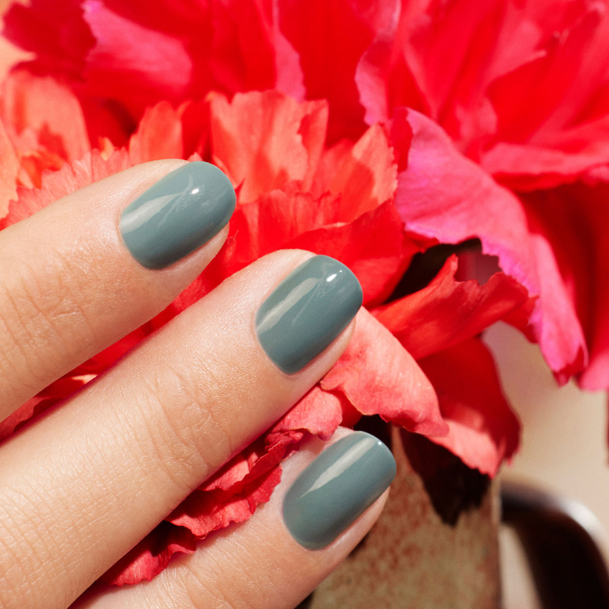 A hand with short, neatly manicured nails painted in glossy Sage by Manucurist is held in front of vibrant pink and red flowers.