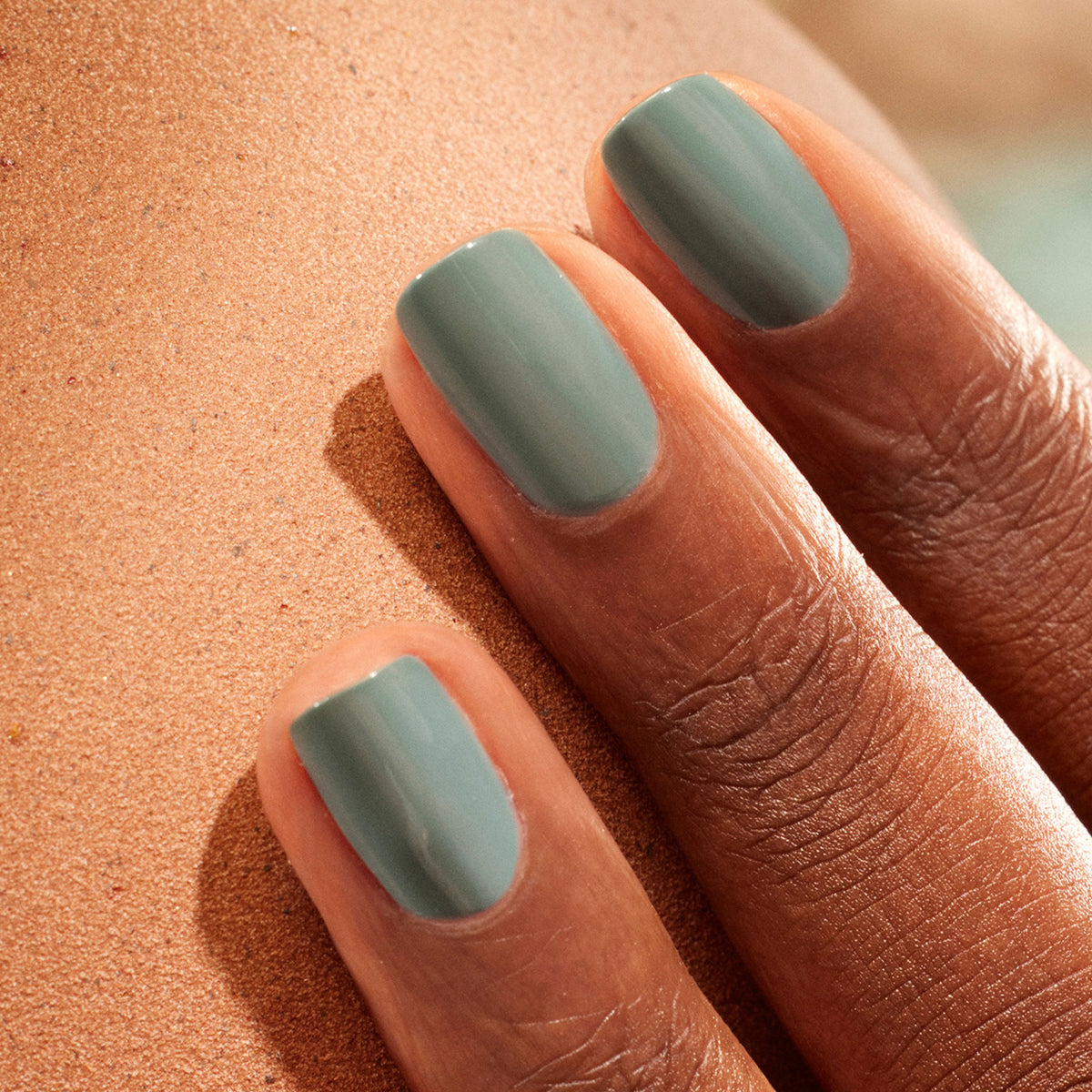 A close-up of three fingers with short, neatly manicured nails painted in Spice Lover by Manucurist, a muted sage green shade, resting against smooth brown skin.