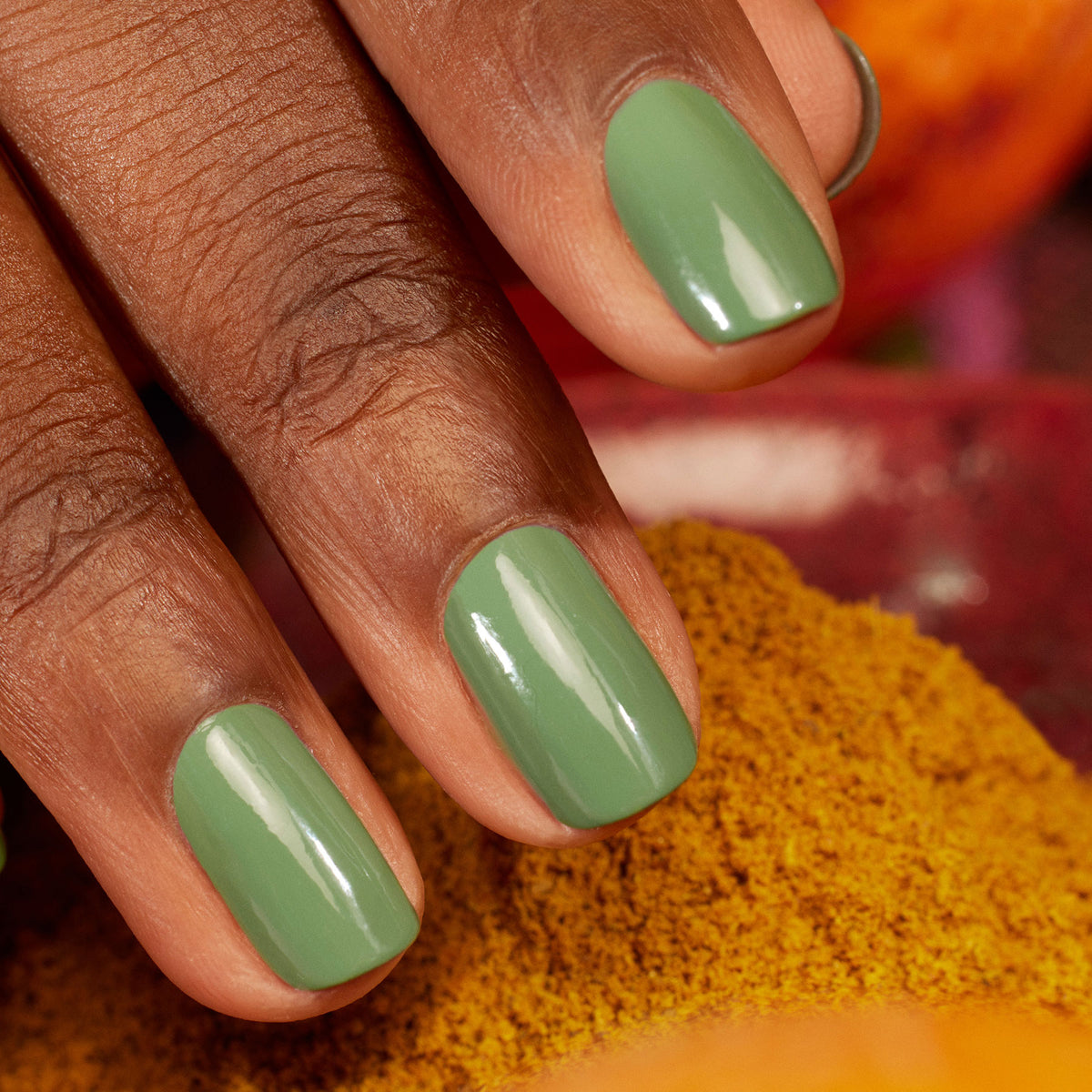 A close-up of a hand with medium brown skin and neatly manicured nails painted in Manucurist Green Flash Artichoke, set against a background of yellow and orange powders.