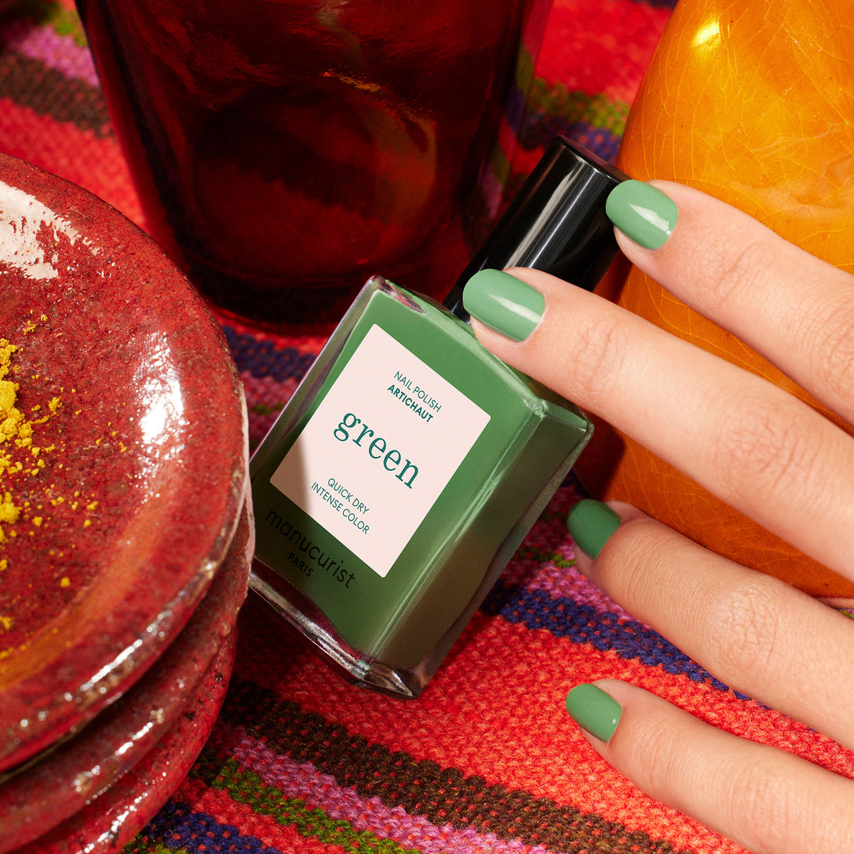 A hand with artichoke green nail polish holds a Manucurist Artichoke bottle, surrounded by colorful dishes and a striped fabric background.