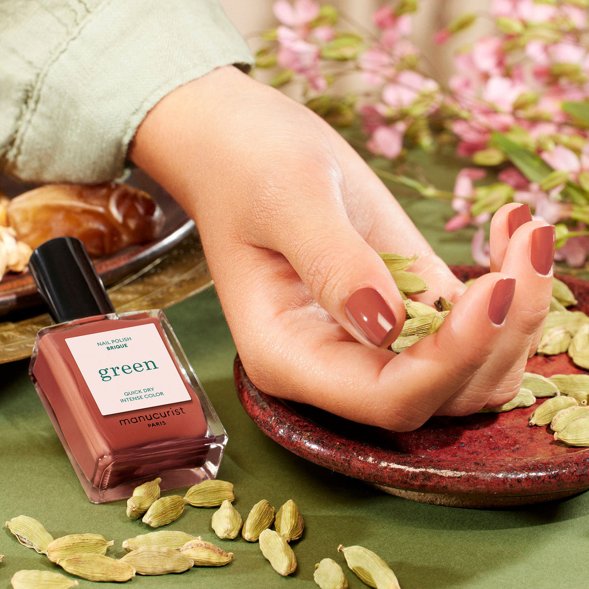 A hand with shiny, brownish-pink Manucurist Brique nail polish rests on a red plate with cardamom pods, evoking the vibrant colors of a Moroccan street market. A Manucurist Brique bottle sits nearby, pink flowers blurred in the background.