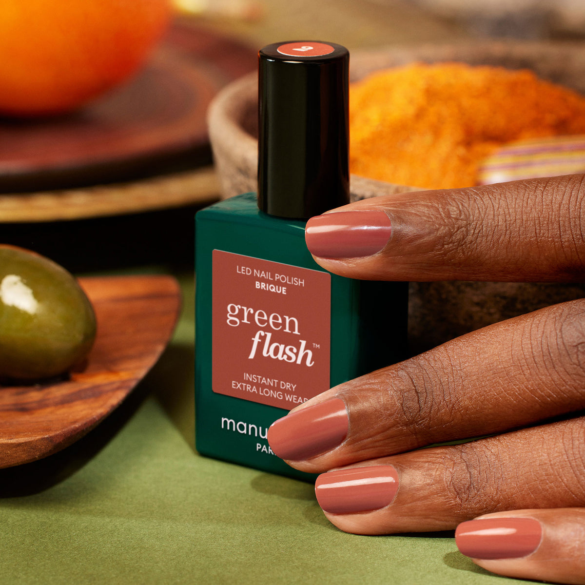 A hand with medium brown skin displays freshly painted nails in Manucurist’s Brique, a muted mauve shade, holding the bottle. In the background are a brick red olive on a wooden dish and a bowl of orange powder.