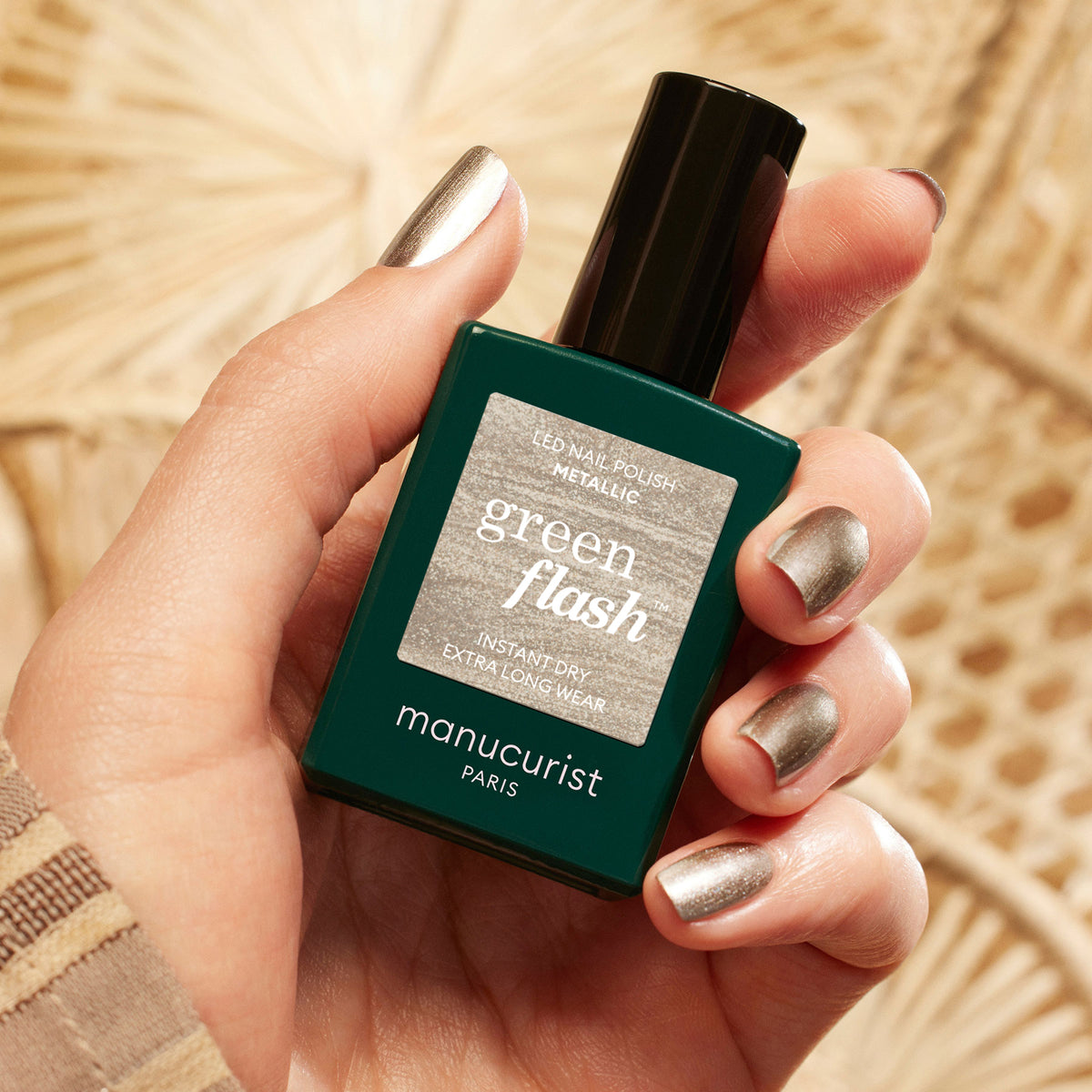 A hand with metallic silver nail polish and highlights holds a bottle of Manucurist Metallic nail polish against a neutral, patterned background.