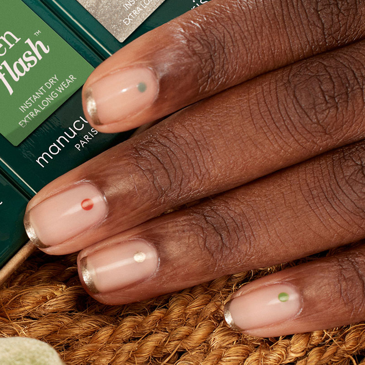 A close-up of a hand with the Manucurist Spice Lover French manicure, each nail displays a different small colored dot near the cuticle. The natural pink base and metallic tips stand out against a textured surface and green Manucurist packaging.