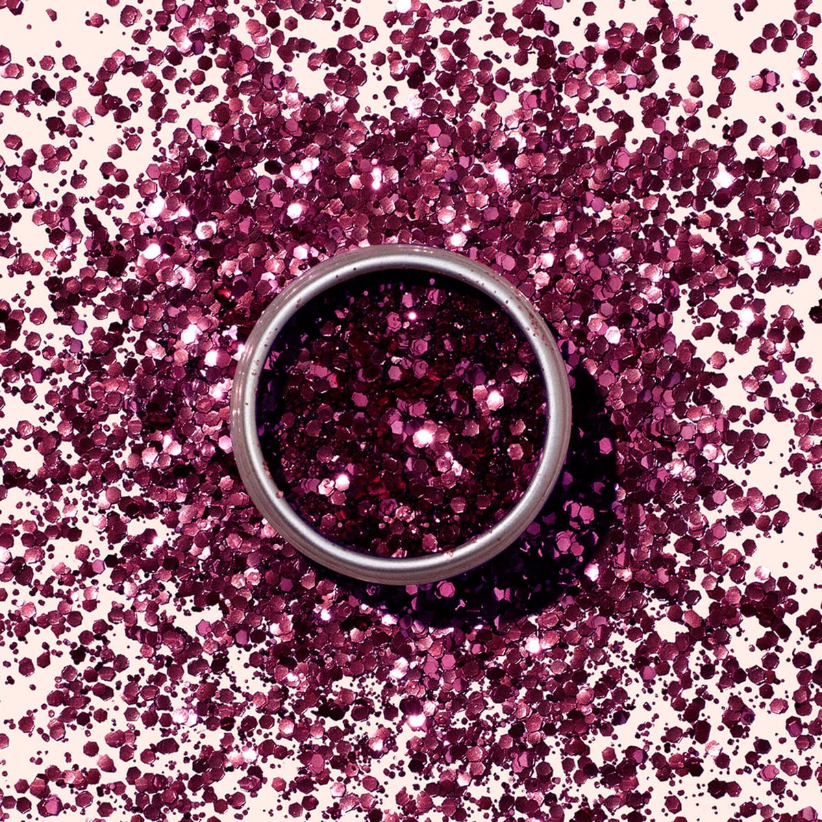 A Manucurist Bordeaux biodegradable glitter surrounds a silver ring on a light surface, forming a sparkling pinkish-purple circular pattern.