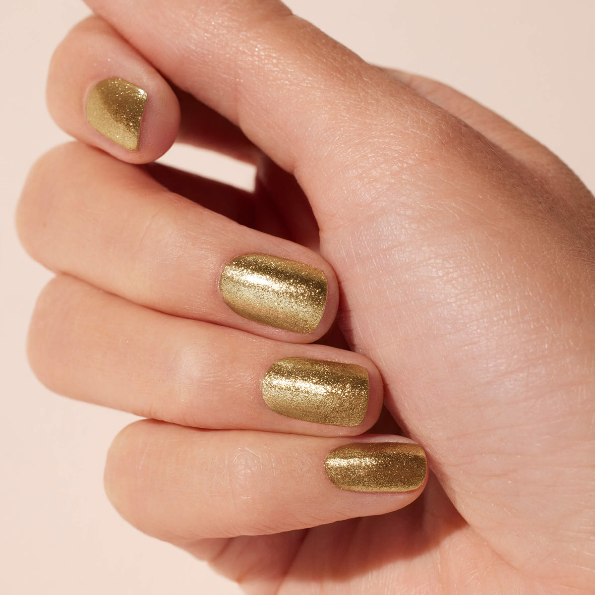 A hand with short, neatly filed nails showcases Manucurist Golden Chrome Powder—shimmery gold gel polish with a subtle chrome finish—against a neutral background.