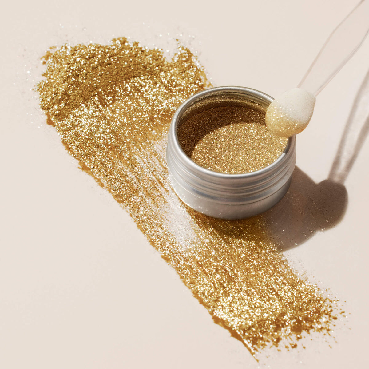 A small open jar of Manucurist Golden Chrome Powder rests on a light surface, its shimmering powder elegantly spread beneath, with a makeup applicator perched on the jar’s edge.