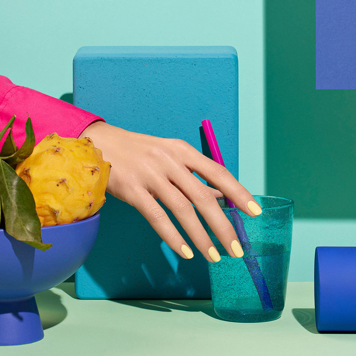 A hand with pastel yellow Smile! nail art by Manucurist holds a turquoise glass with a pink straw, next to a blue bowl of yellow fruit and greens, all set on a vibrant blue and turquoise geometric Yucatán collection background.