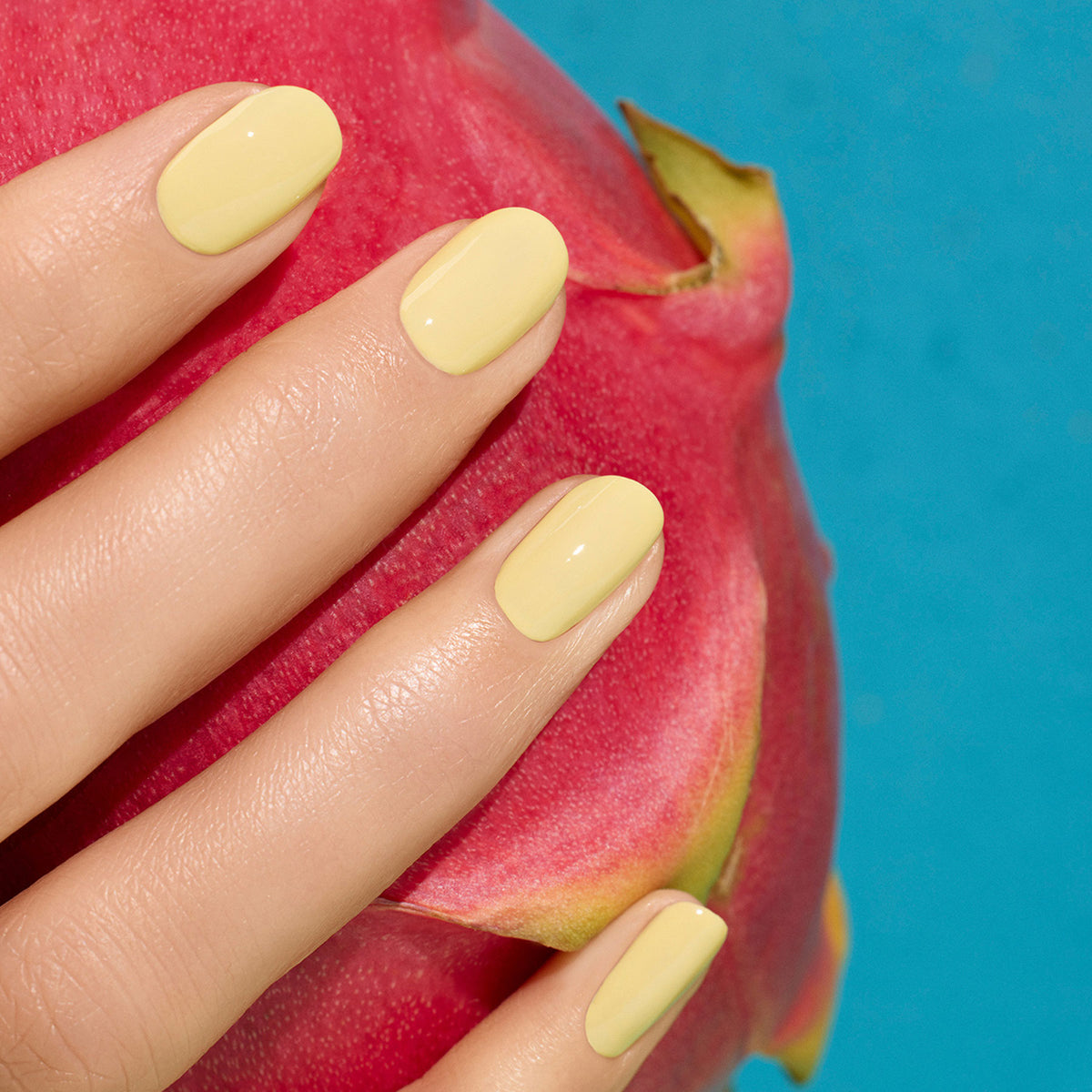 A hand with smooth, yellow-painted nails in Manucurist’s Flower love shade rests on a pink dragon fruit against a bright blue background.