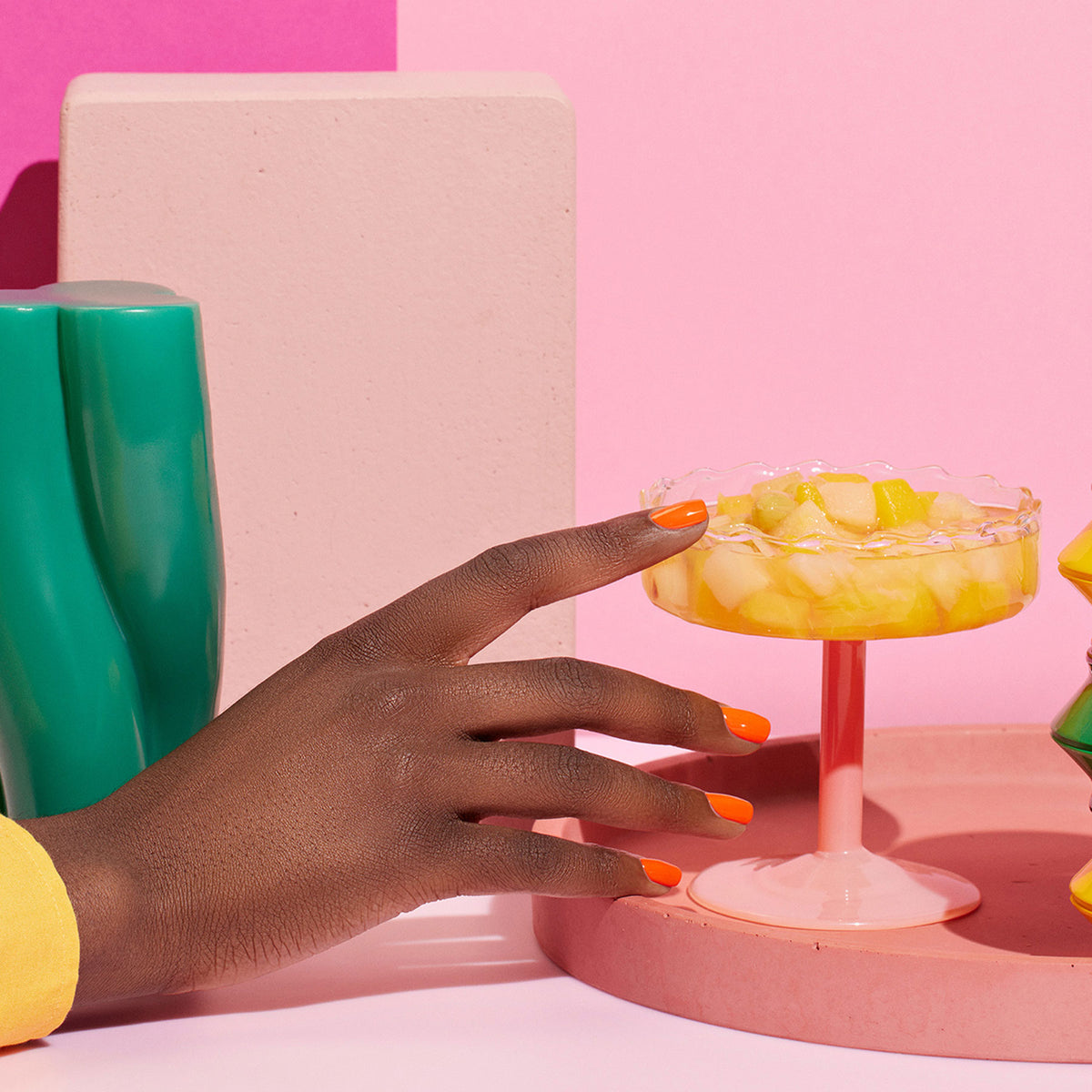 A hand with Smile! by Manucurist orange nails reaches for a pink glass dessert dish of fruit salad. The green, pink, and beige geometric backdrop offers summer nail art inspiration from the Yucatán collection.