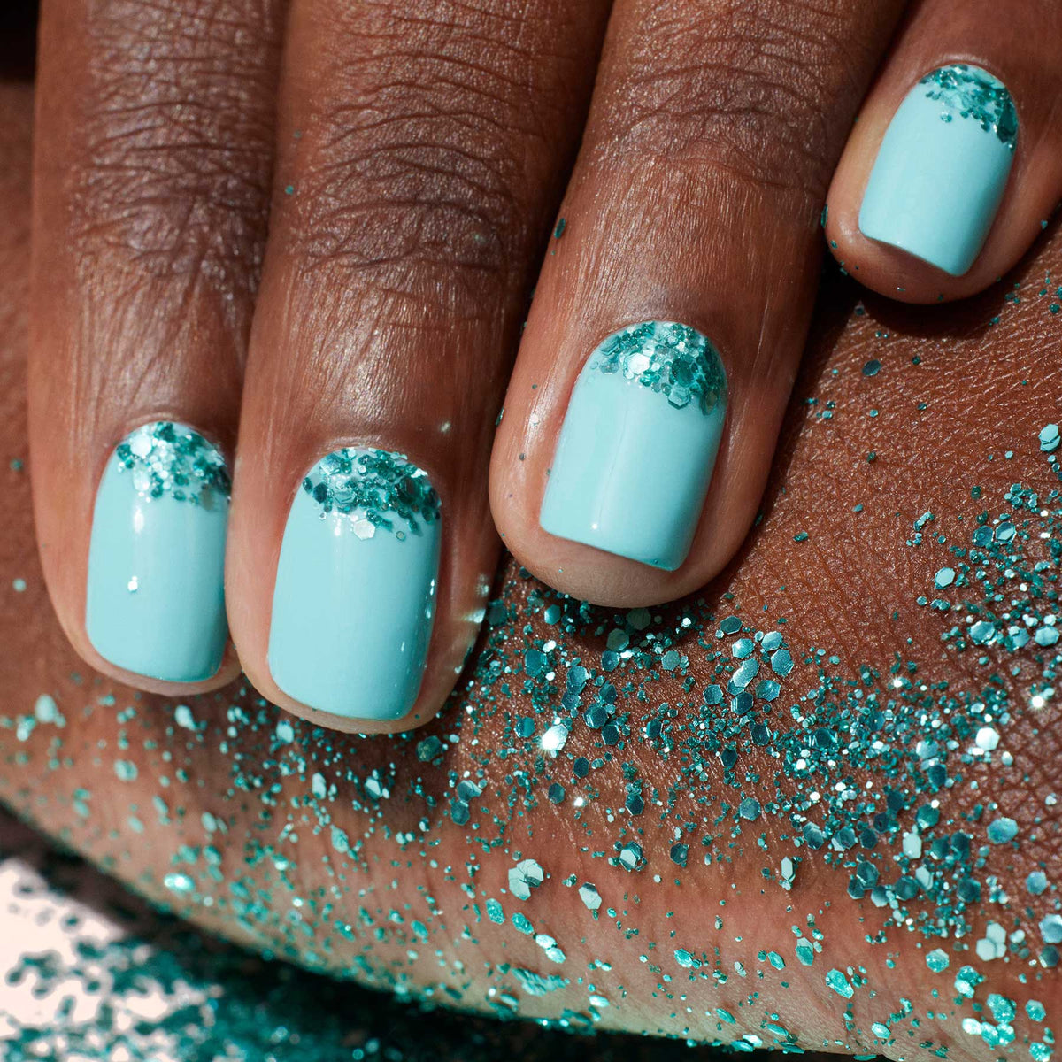 A hand with short, neatly manicured nails painted in light blue and detailed using a fine nail art brush, features Manucurist’s Underwater glitter—biodegradable, turquoise glitter near each cuticle—resting on skin with matching sparkle.